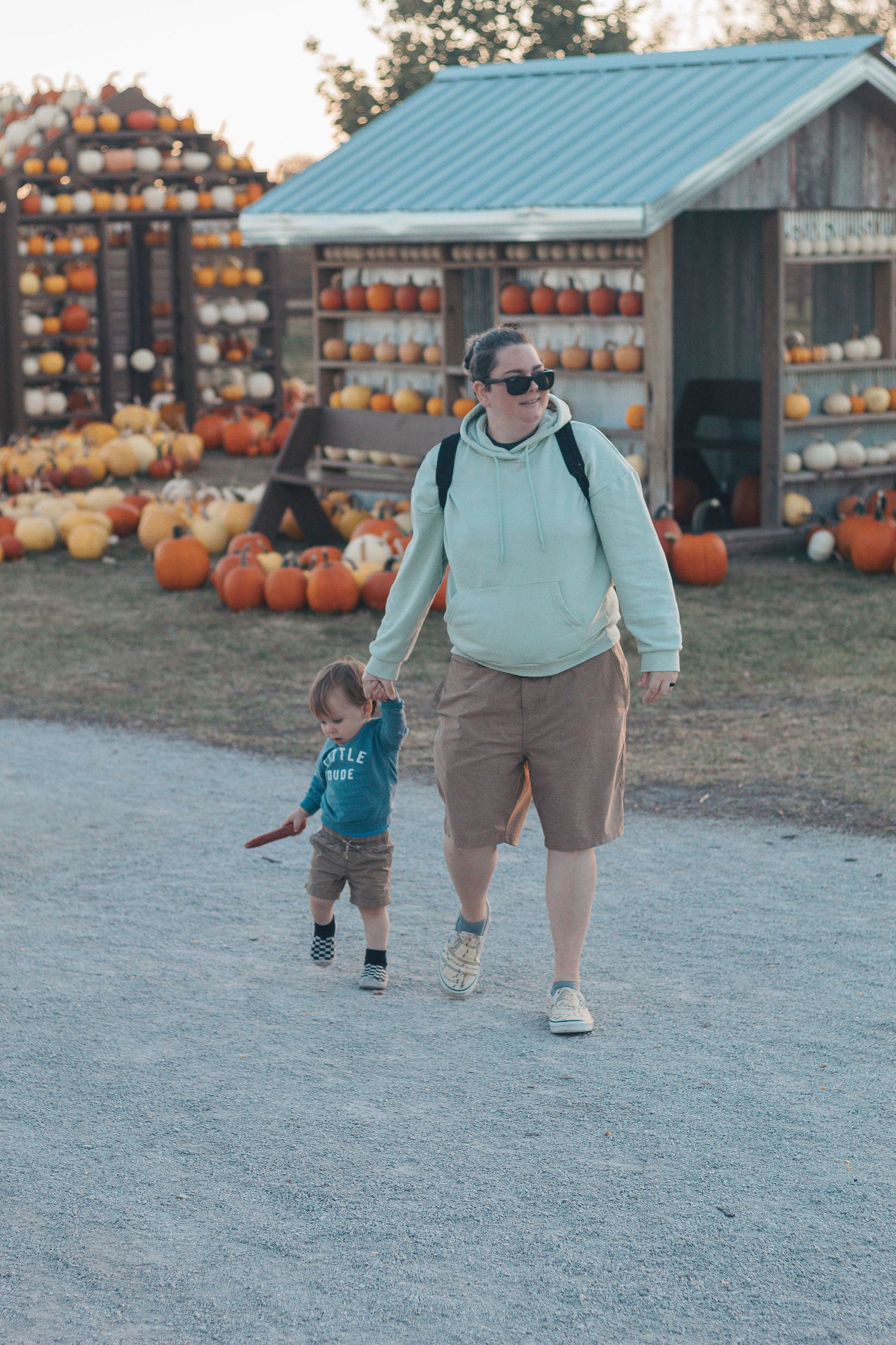 Two people walking through a pumpkin patch with rustic wooden barns and orange pumpkins displayed in the background.