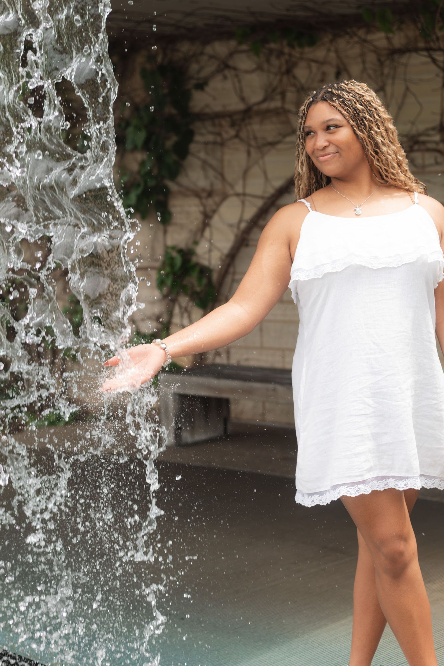 Young woman in white dress touching a cascading waterfall fountain outdoors, smiling joyfully.