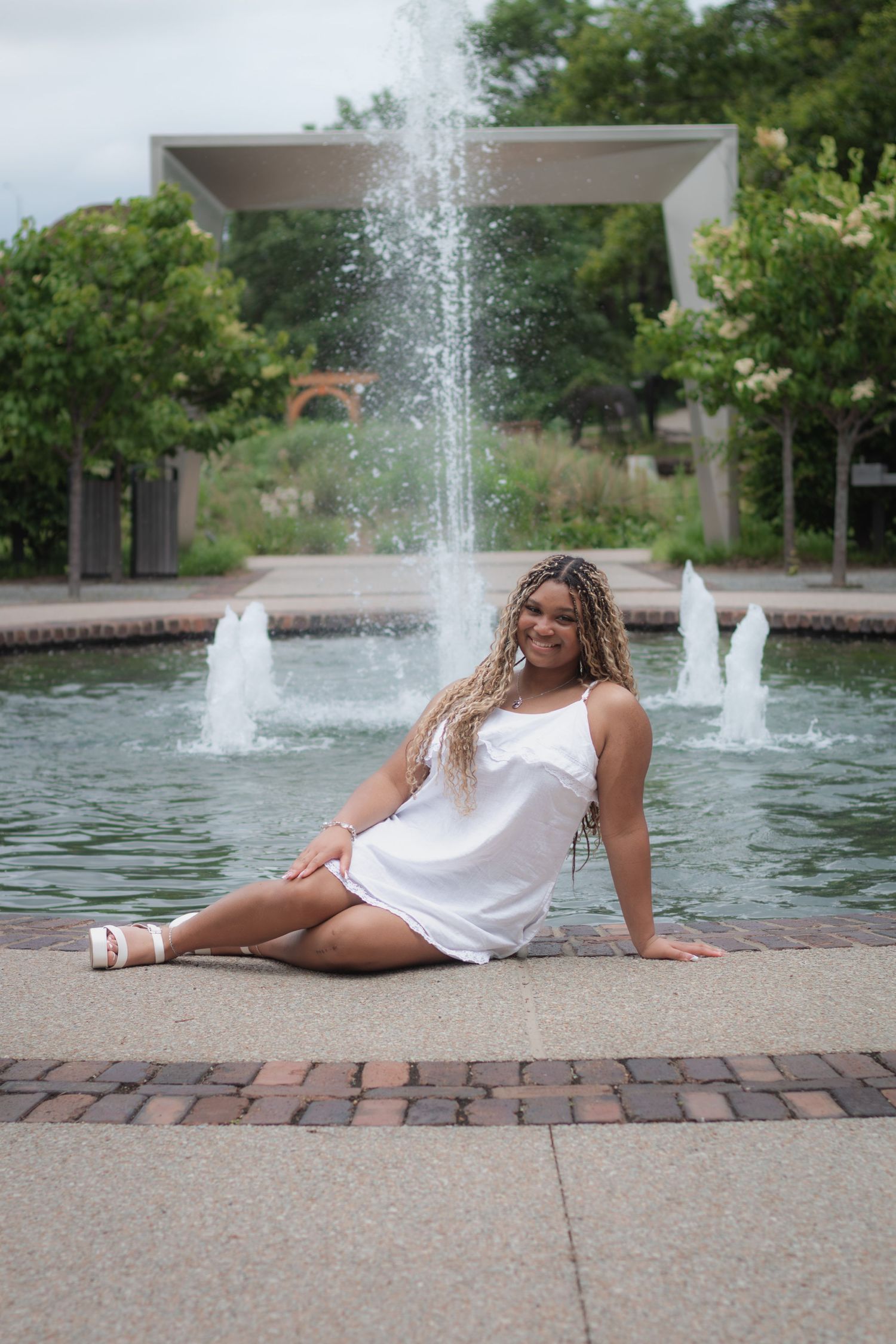 Young woman in white dress sitting gracefully by a decorative fountain in a lush green park, smiling confidently.
