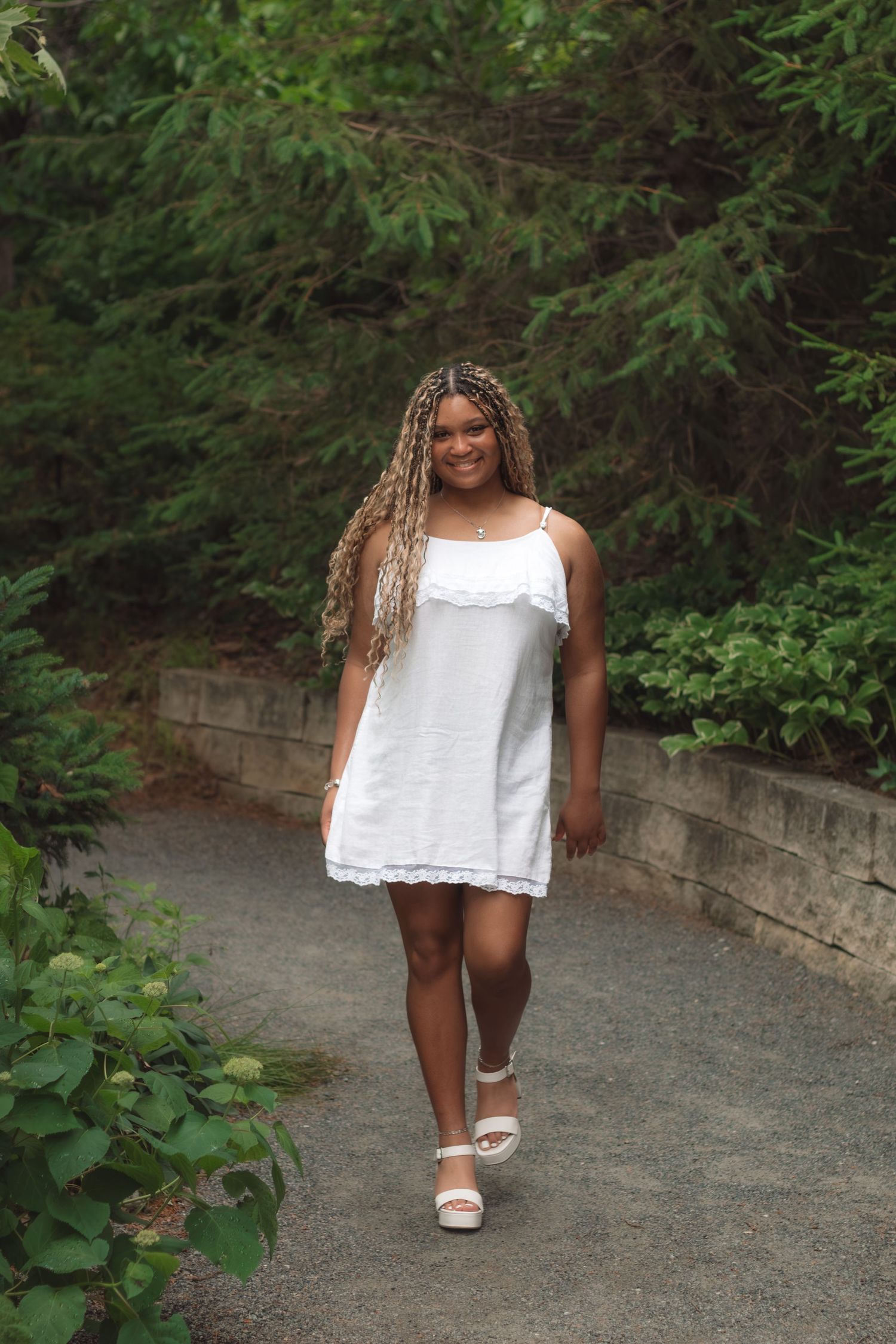Young girl in white sundress with long braided hair walks on a garden path surrounded by lush green trees.