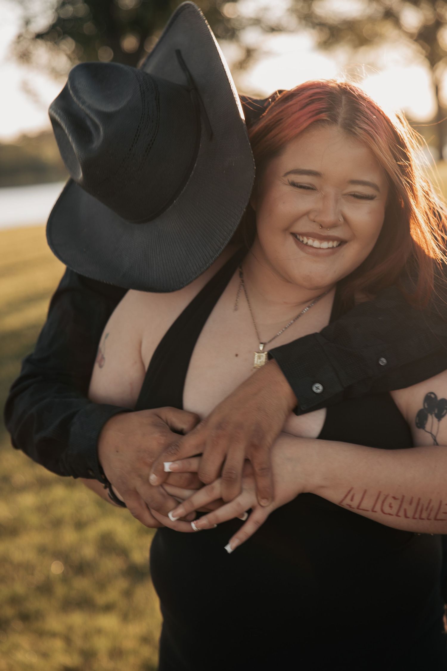 A smiling redhead woman with tattoos is embraced from behind by a partner wearing a black cowboy hat outdoors.