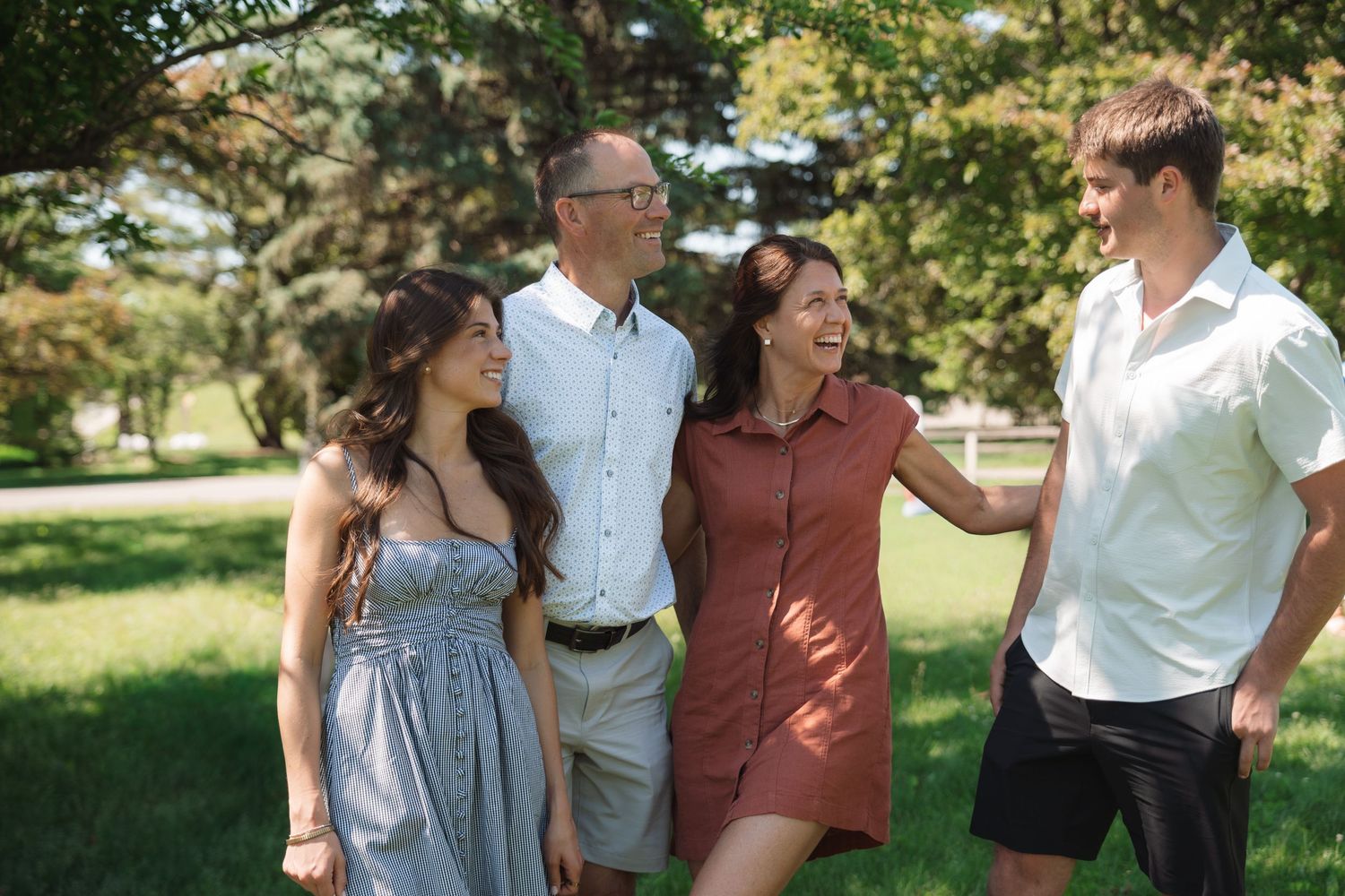 Family of four smiling and walking together in a sunny park surrounded by lush green trees.