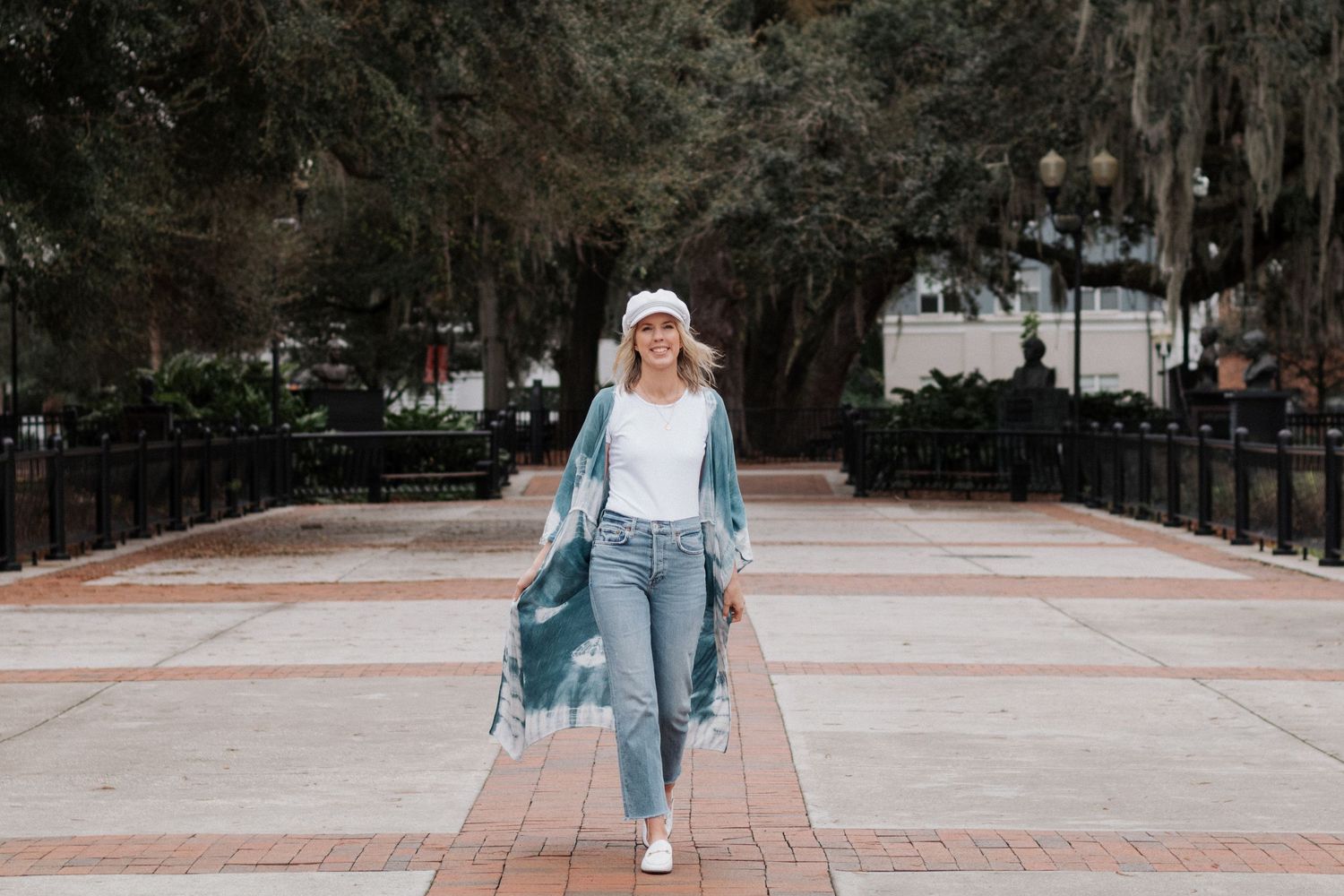 A person wearing a long blue cardigan and ripped jeans walks on a brick paved path surrounded by trees.