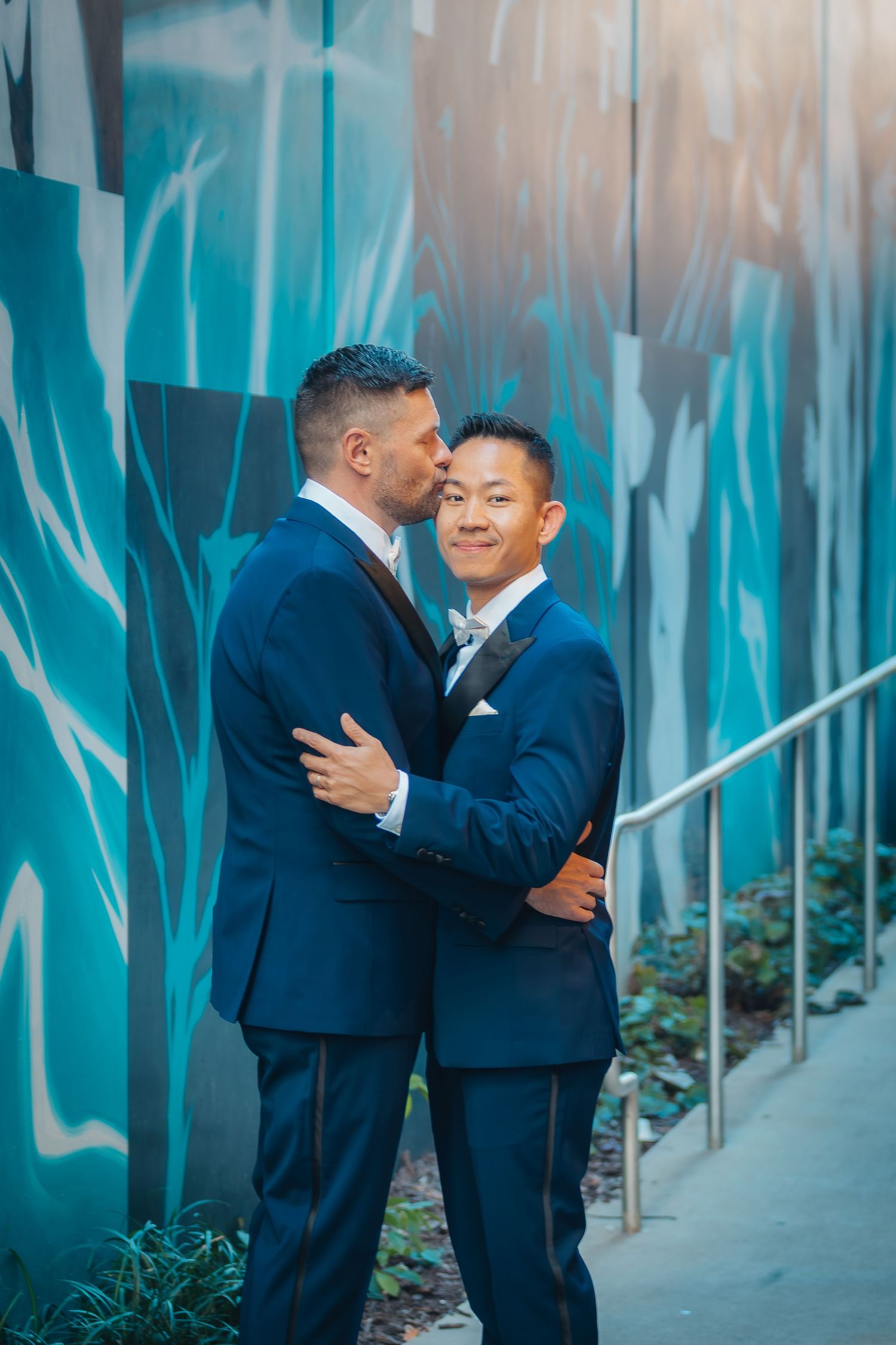 Two people in navy blue suits embrace in front of a turquoise wall with modern architectural details.