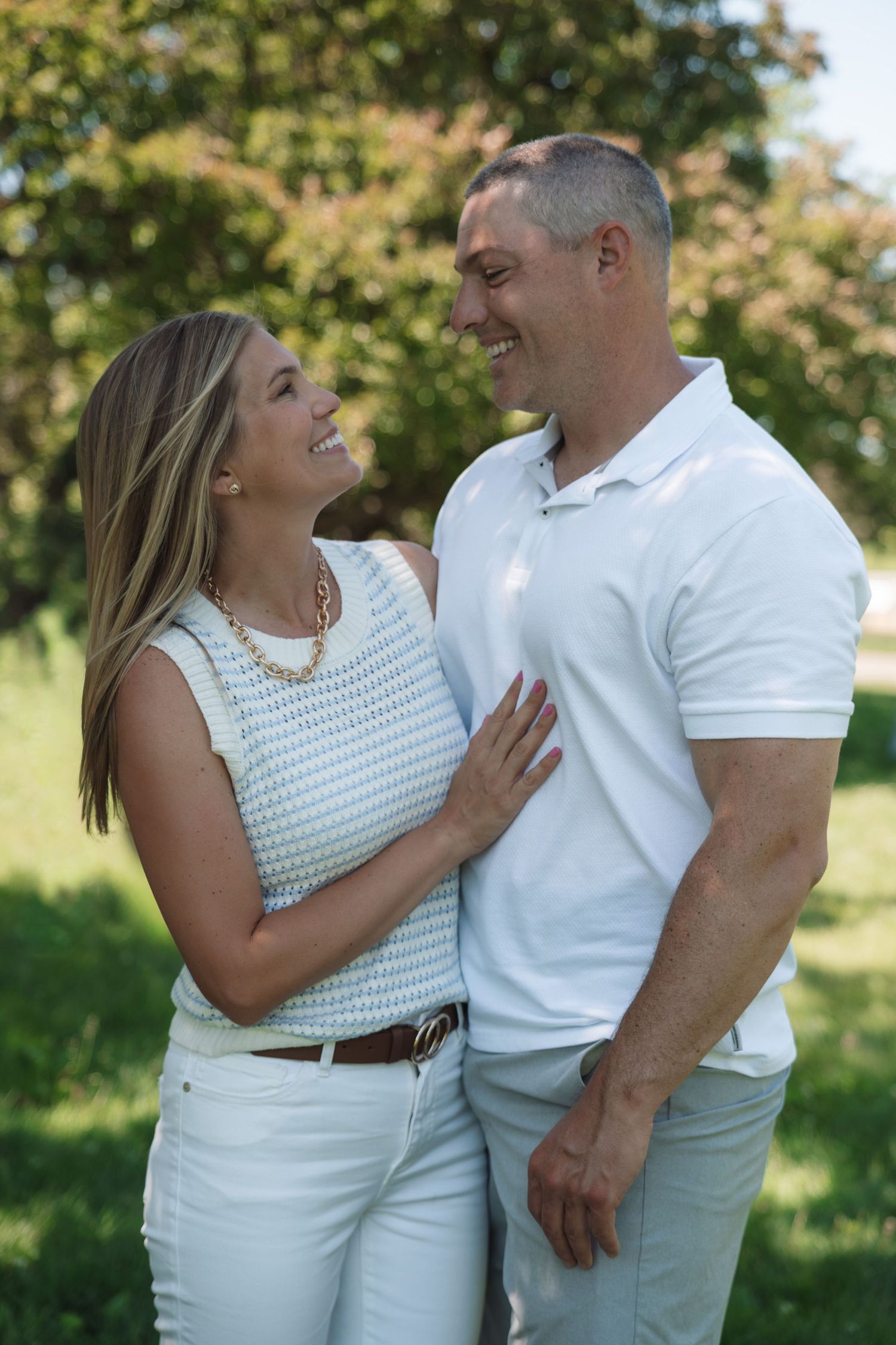 A smiling couple in matching white outfits share a joyful moment outdoors in a lush green park setting.