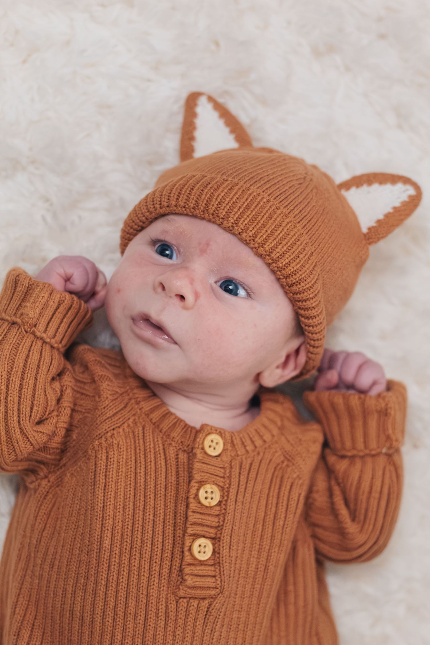 Baby wearing a cozy brown knit sweater and matching hat lies on soft cream fabric.