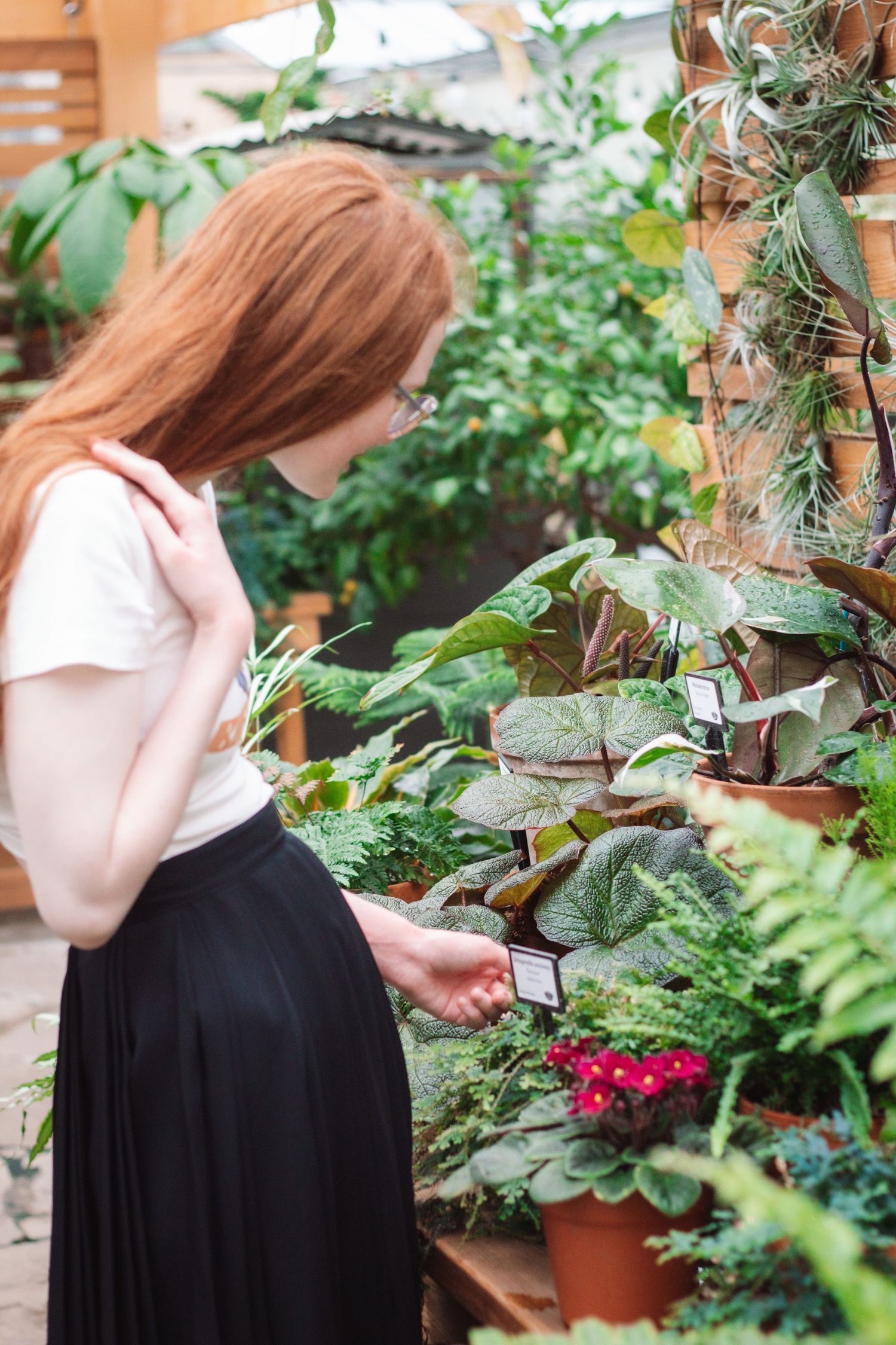 A young redhead woman in a white top and black skirt browses potted plants at a garden shop.