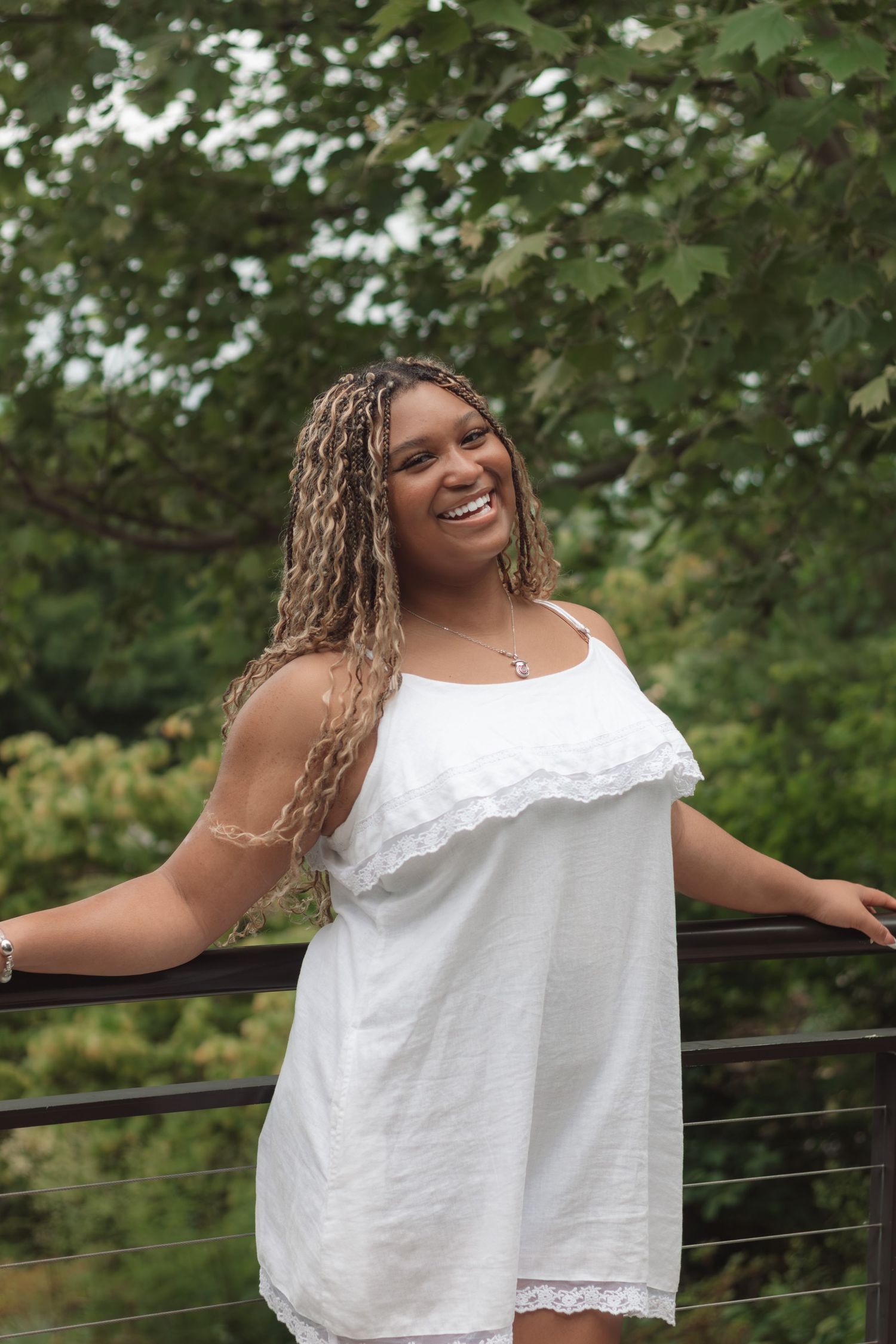 Smiling young woman with braided hair wearing a white sundress, standing on a bridge surrounded by lush green trees.