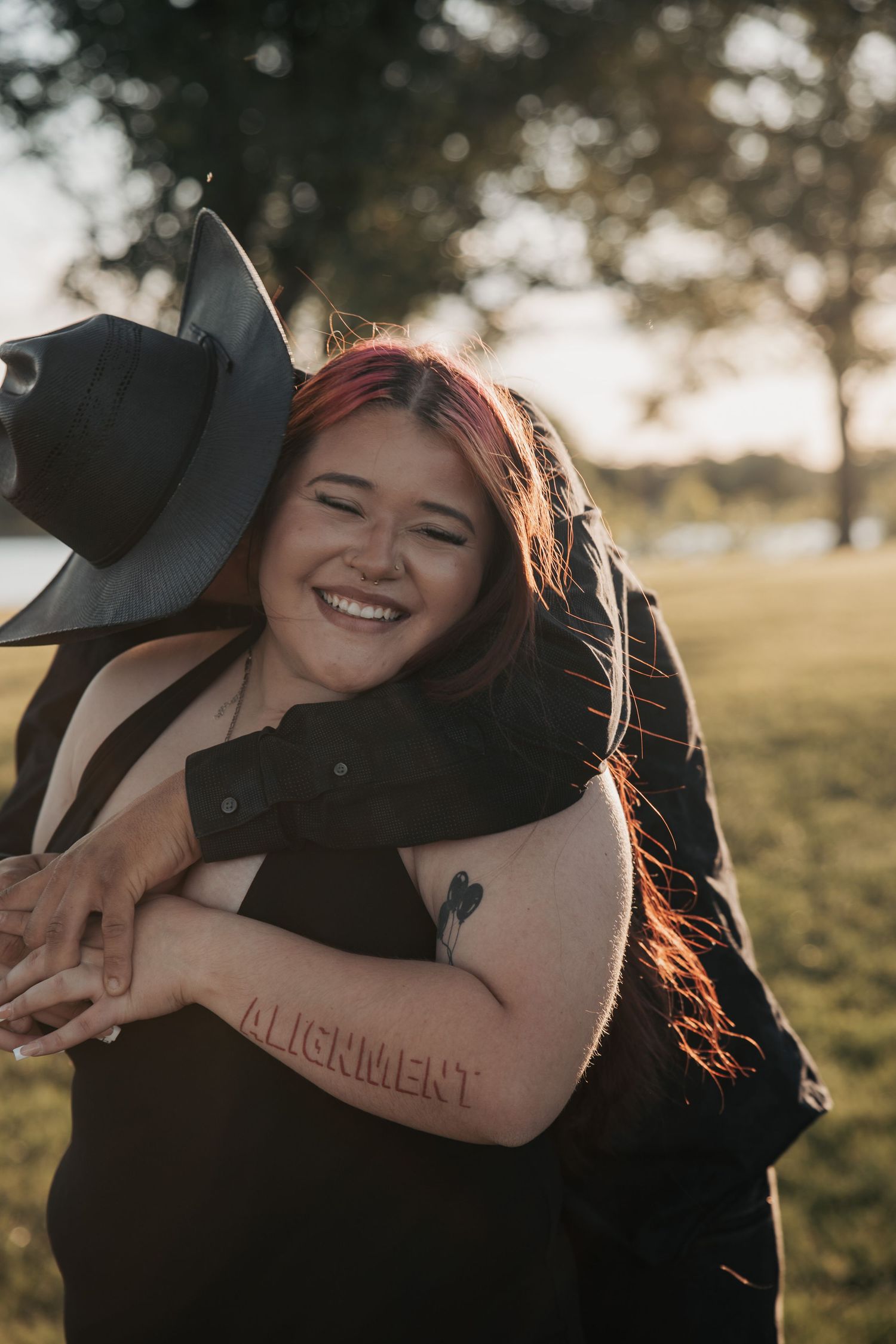 A smiling woman with red hair and an ALIGNMENT tattoo hugs a person in a black cowboy hat outdoors at golden hour.