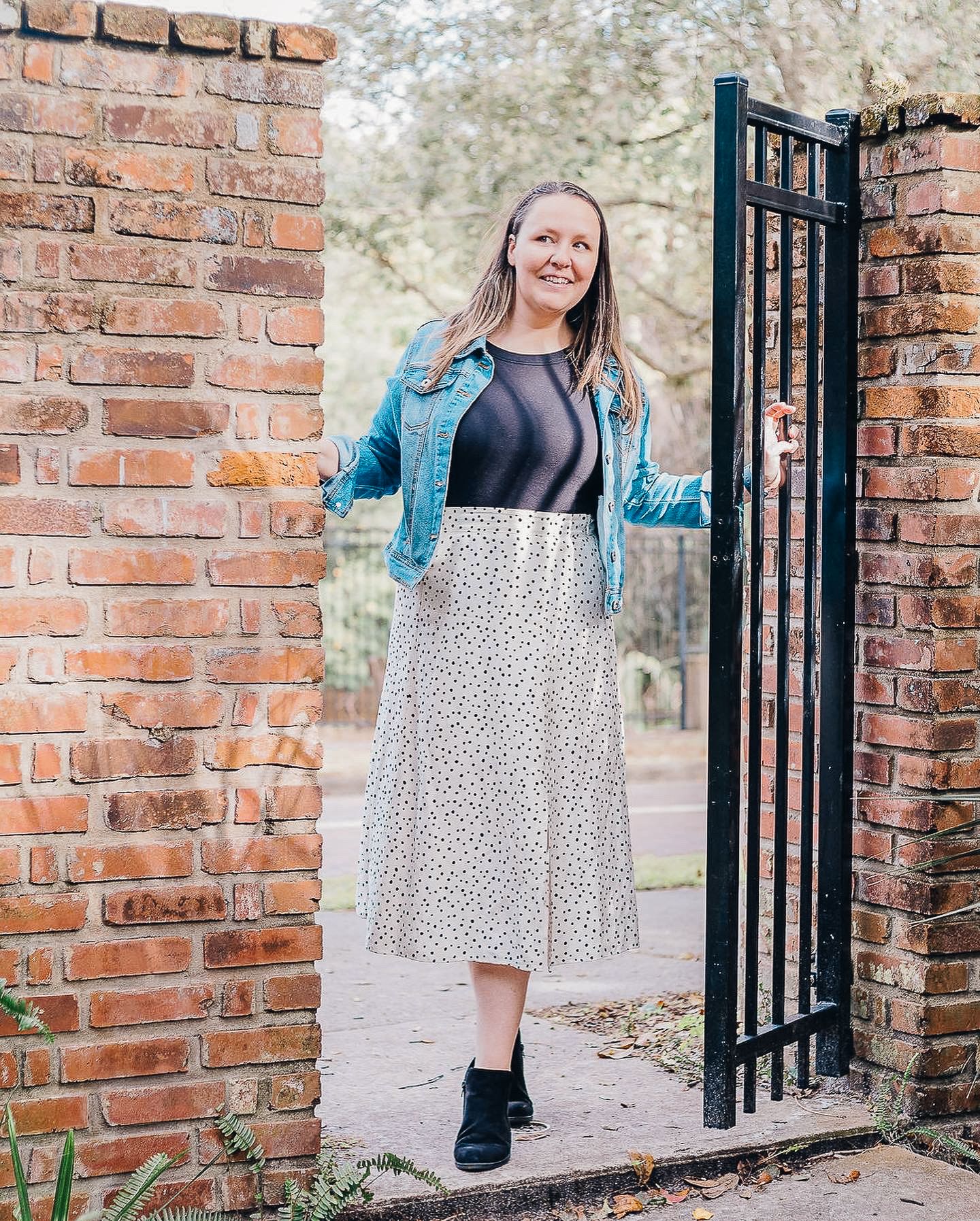 Someone wearing a denim jacket and spotted midi skirt stands smiling in a brick doorway entrance.