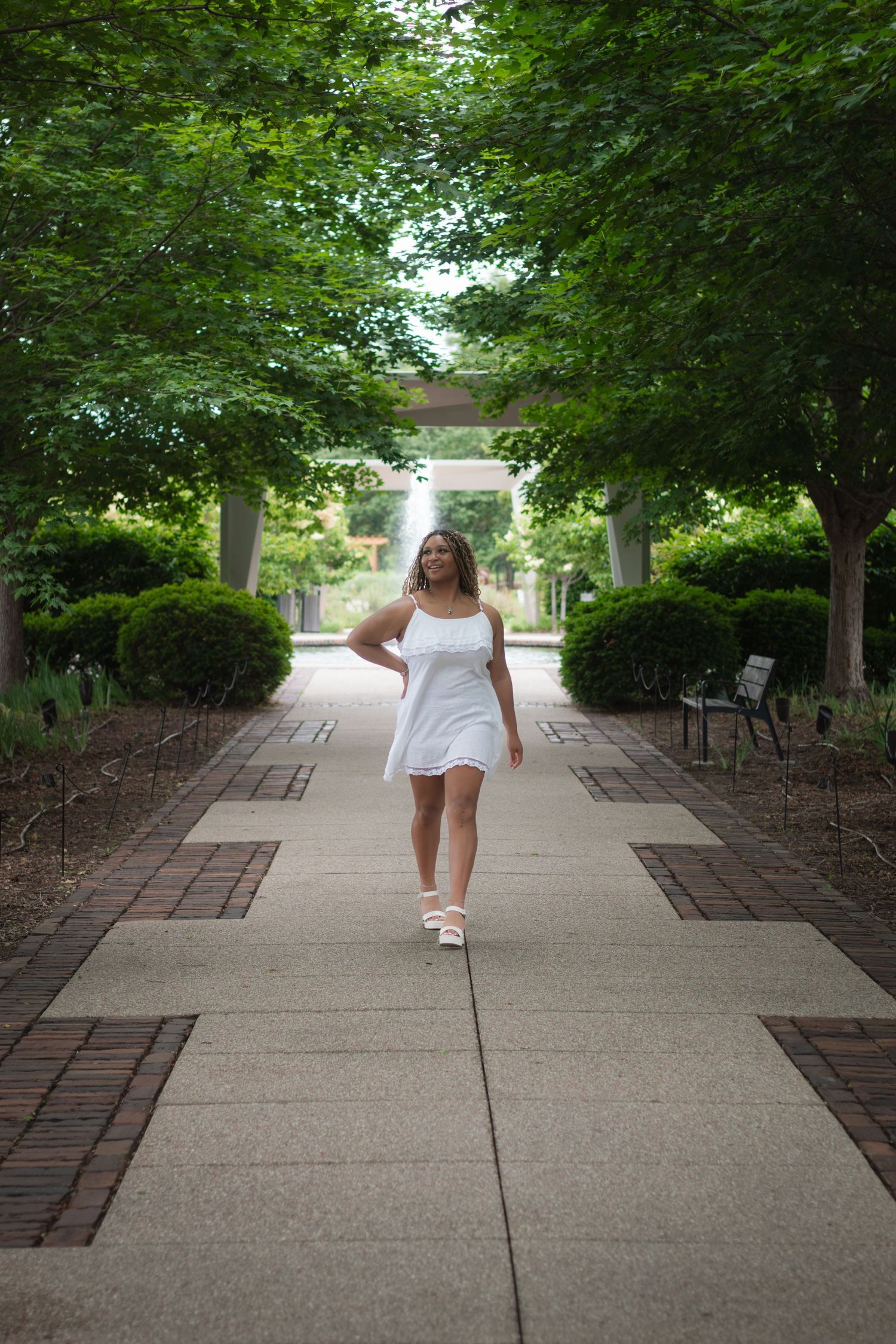 Woman in white dress walking on a tree-lined garden path with lush green foliage and a fountain backdrop.