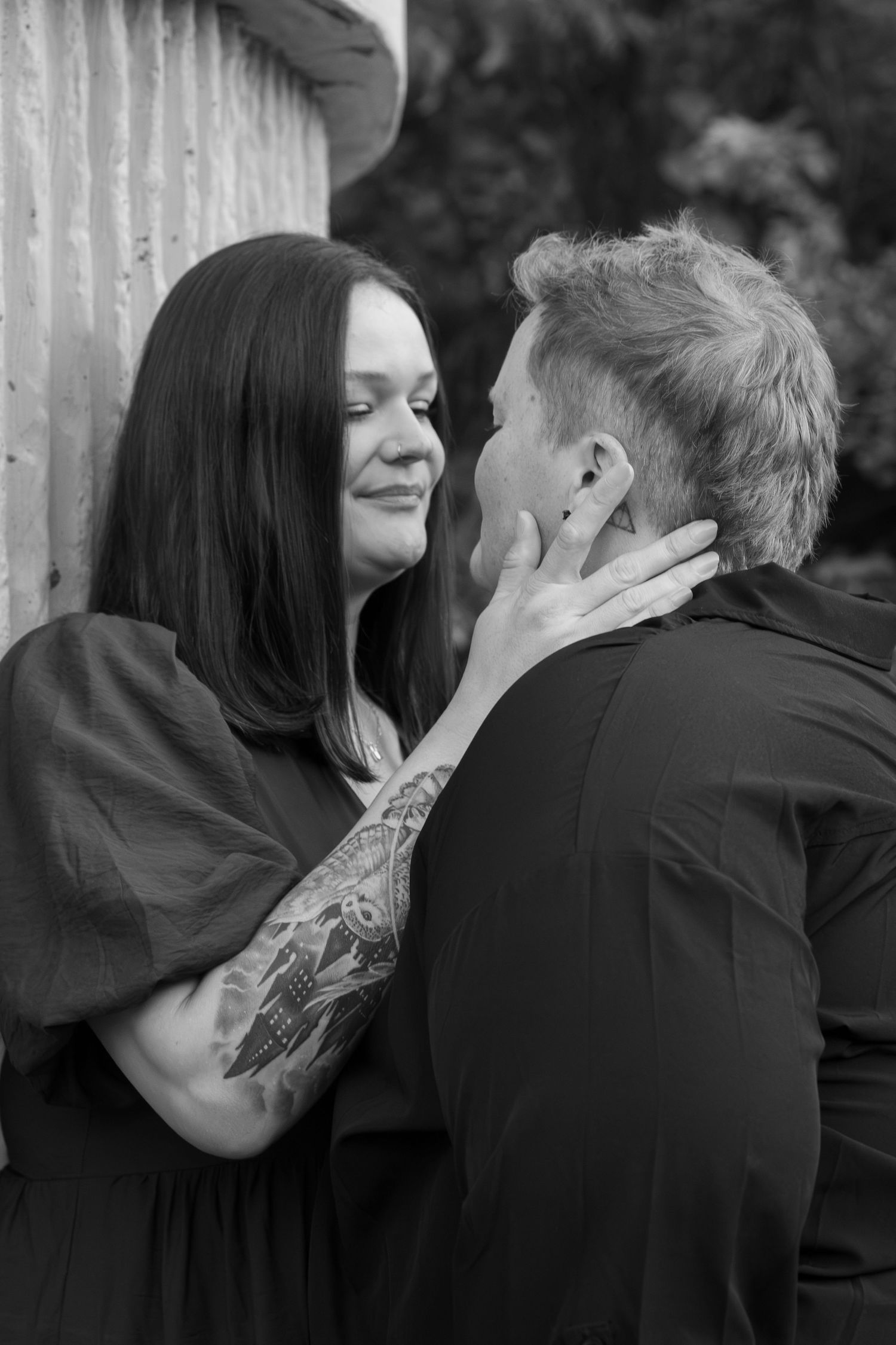 Black and white photo of a tattooed woman tenderly holding a man's face as they share an intimate moment outdoors.