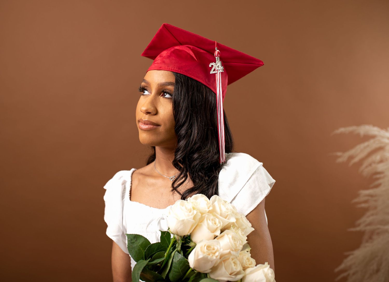 A graduate in a red cap and white gown holds white roses against a brown backdrop for their portrait.