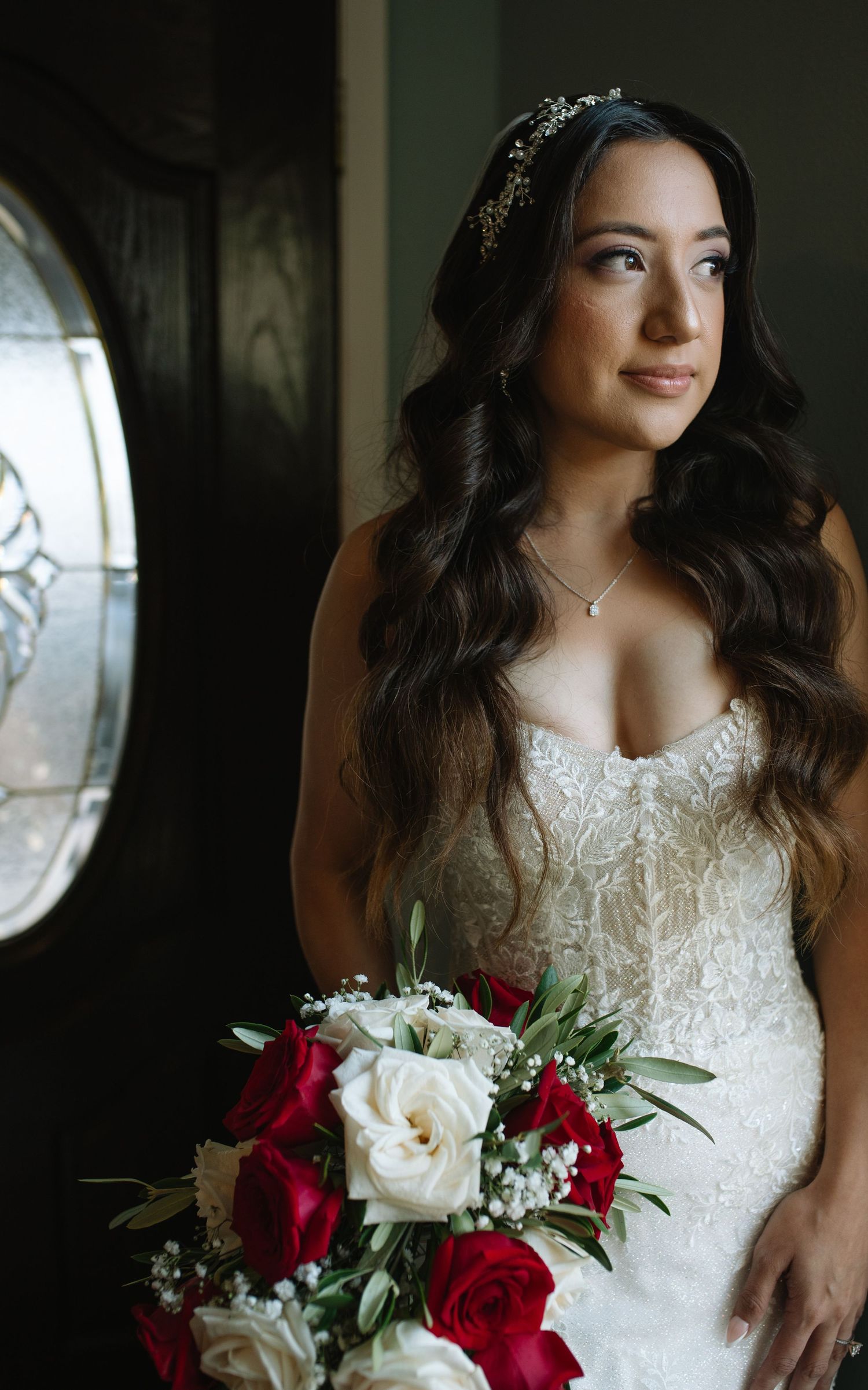 A bride in a white lace wedding dress holds a stunning bouquet of red and white roses against a dark background.