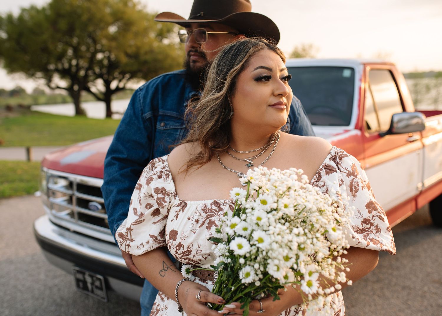Couple in floral lace dress holds white baby's breath bouquet with vintage red pickup truck in background.