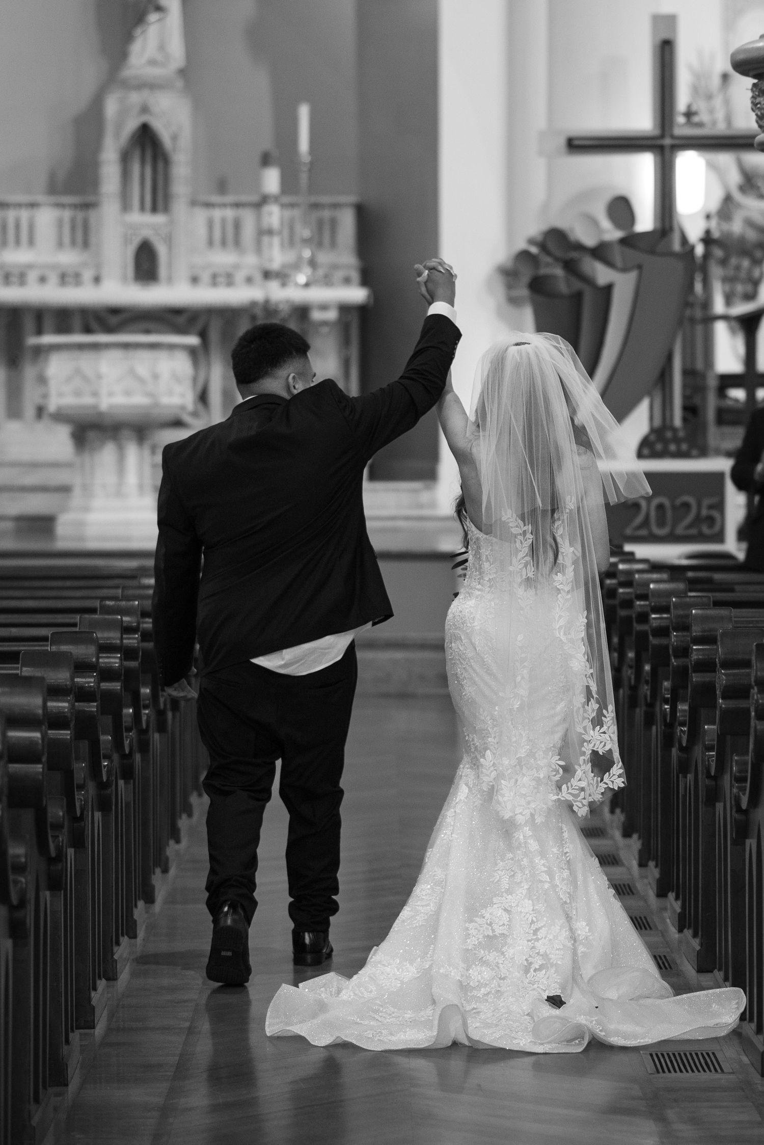 A newlywed couple celebrates with raised hands while walking down the church aisle after their wedding ceremony.
