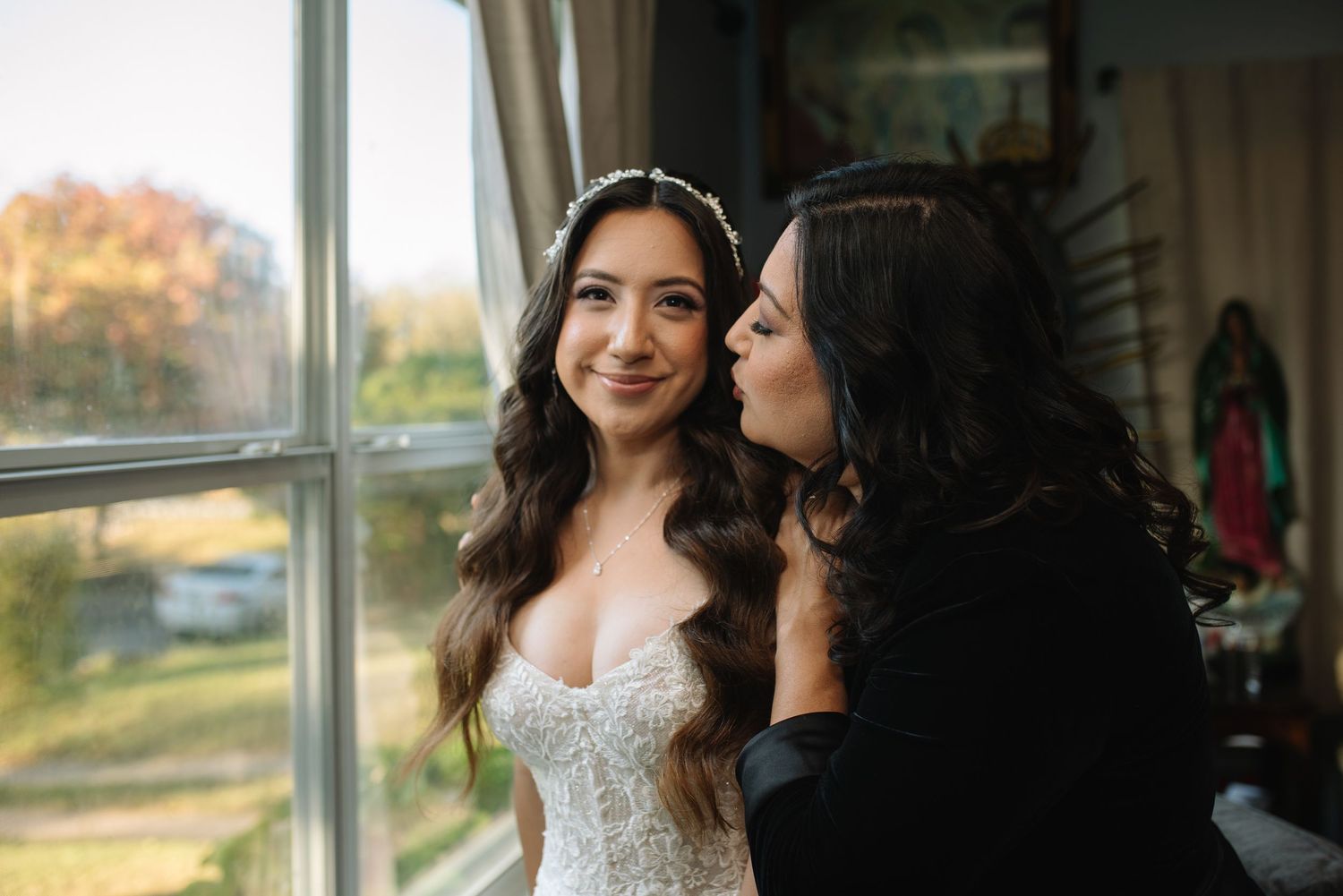 A bride and braids mom sharing a tender moment while getting ready in a sunlit room before the wedding ceremony.