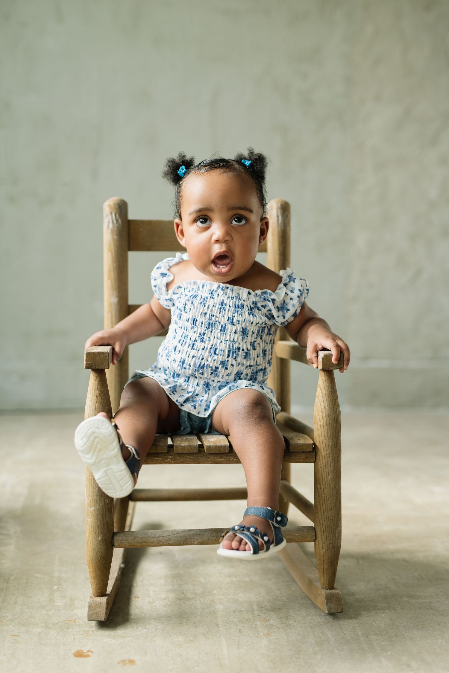 Adorable baby girl sitting in a wooden rocking chair, wearing a blue floral romper and sandals, with a surprised expression.