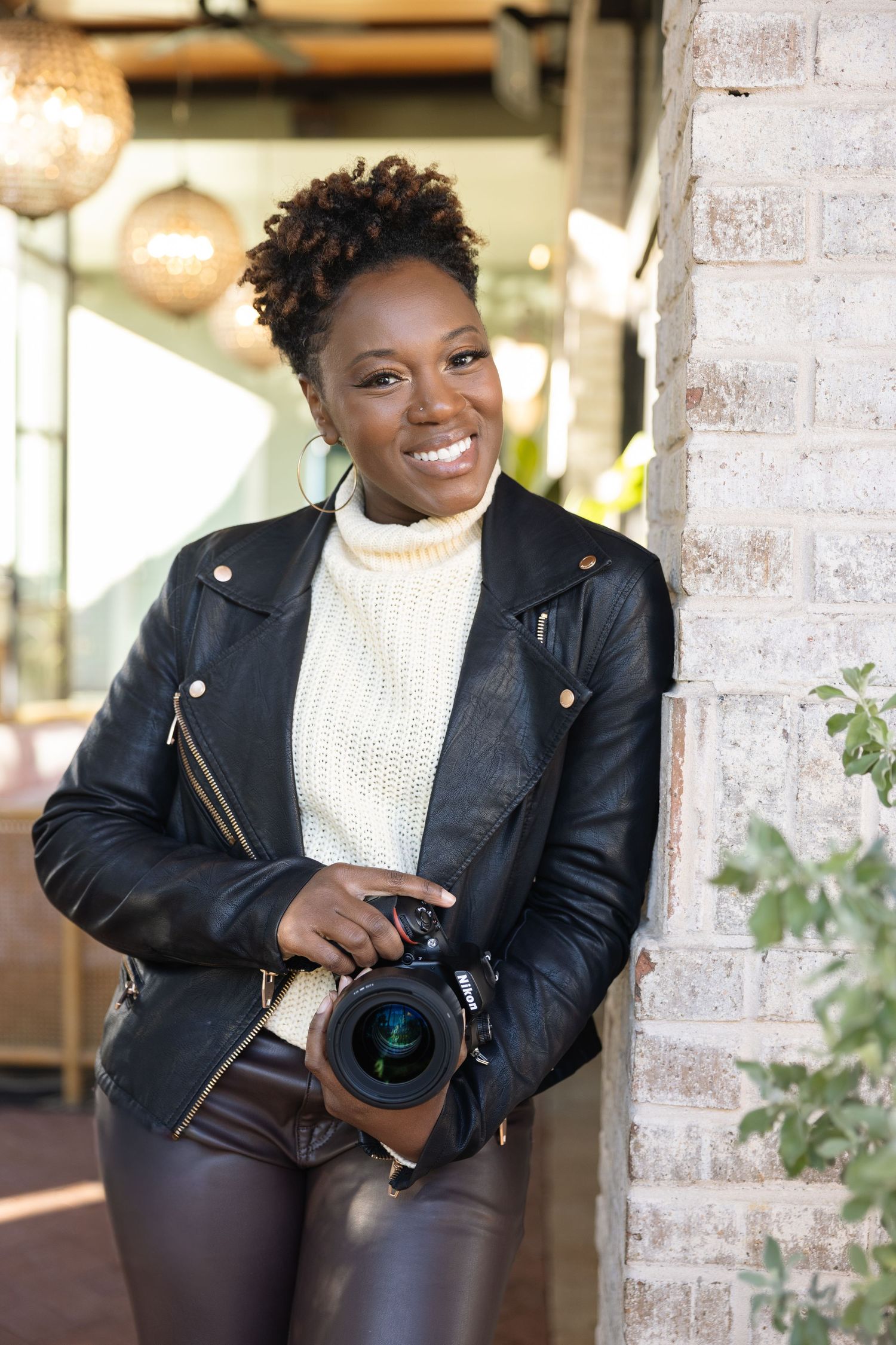 Professional photographer in leather jacket and white sweater poses with camera against brick wall in studio setting.