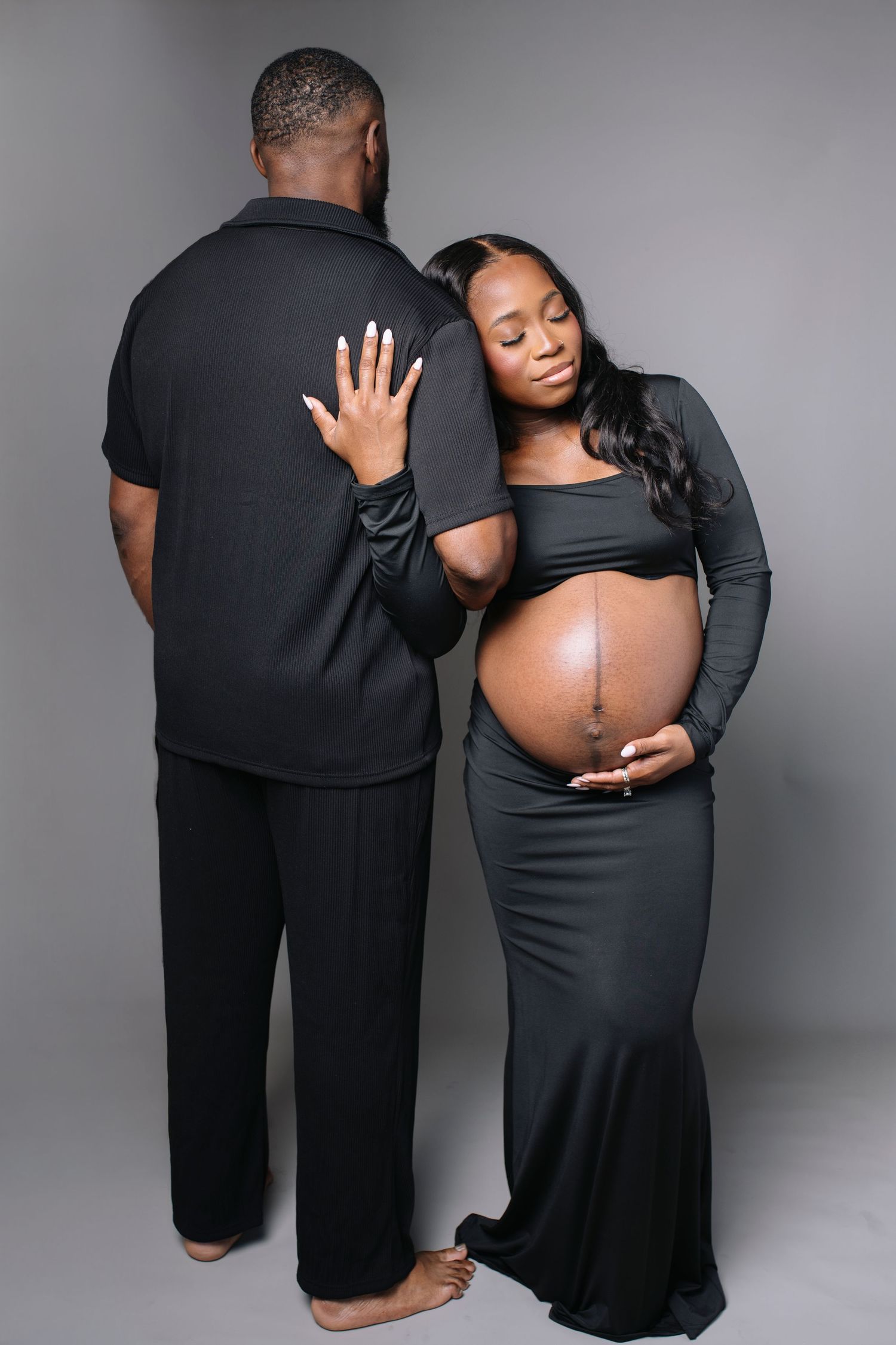 A series of intimate maternity photos showing a couple in black clothing embracing against a grey studio backdrop.