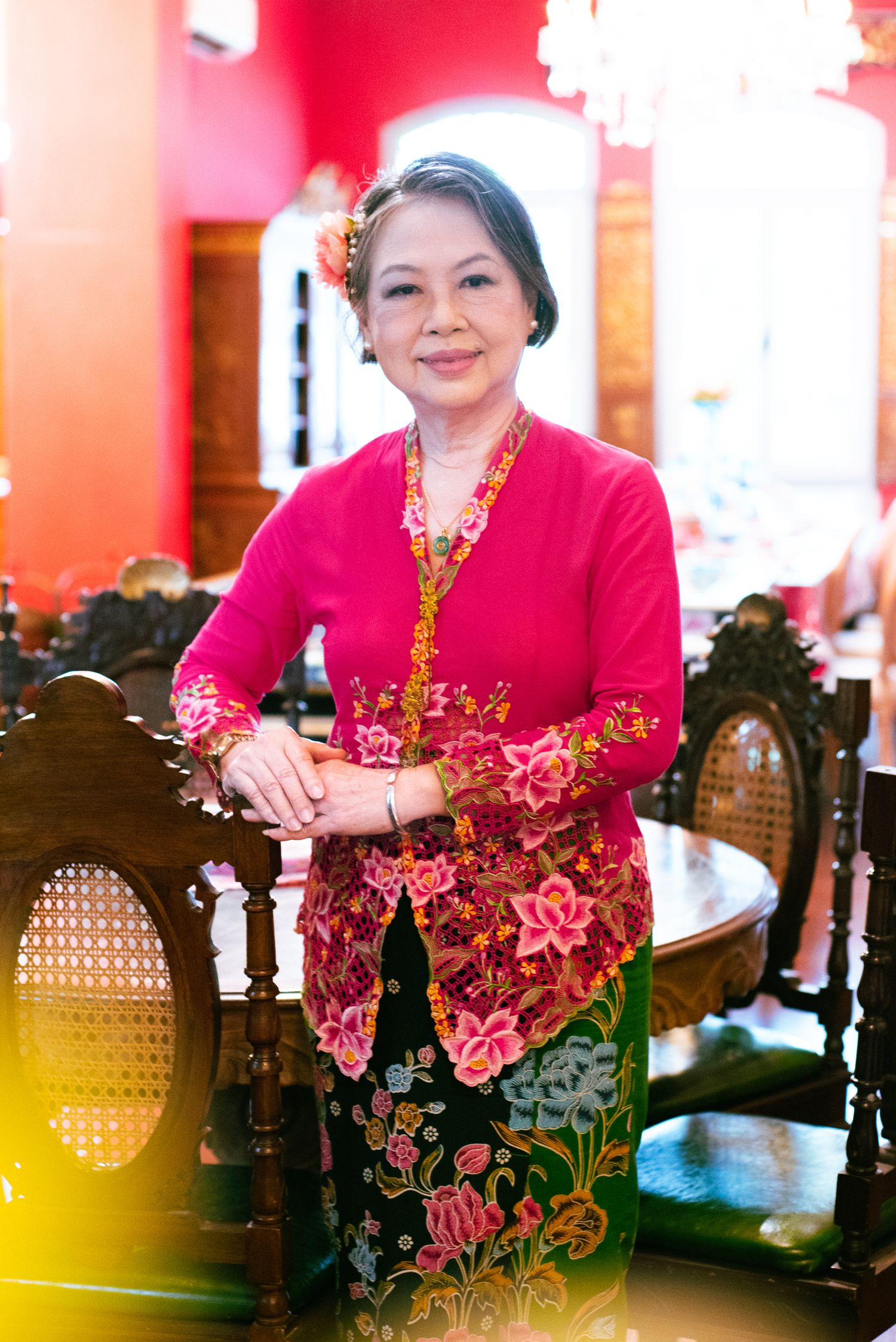 Traditional Indonesian kebaya outfit in red and pink floral patterns photographed in warm interior setting.