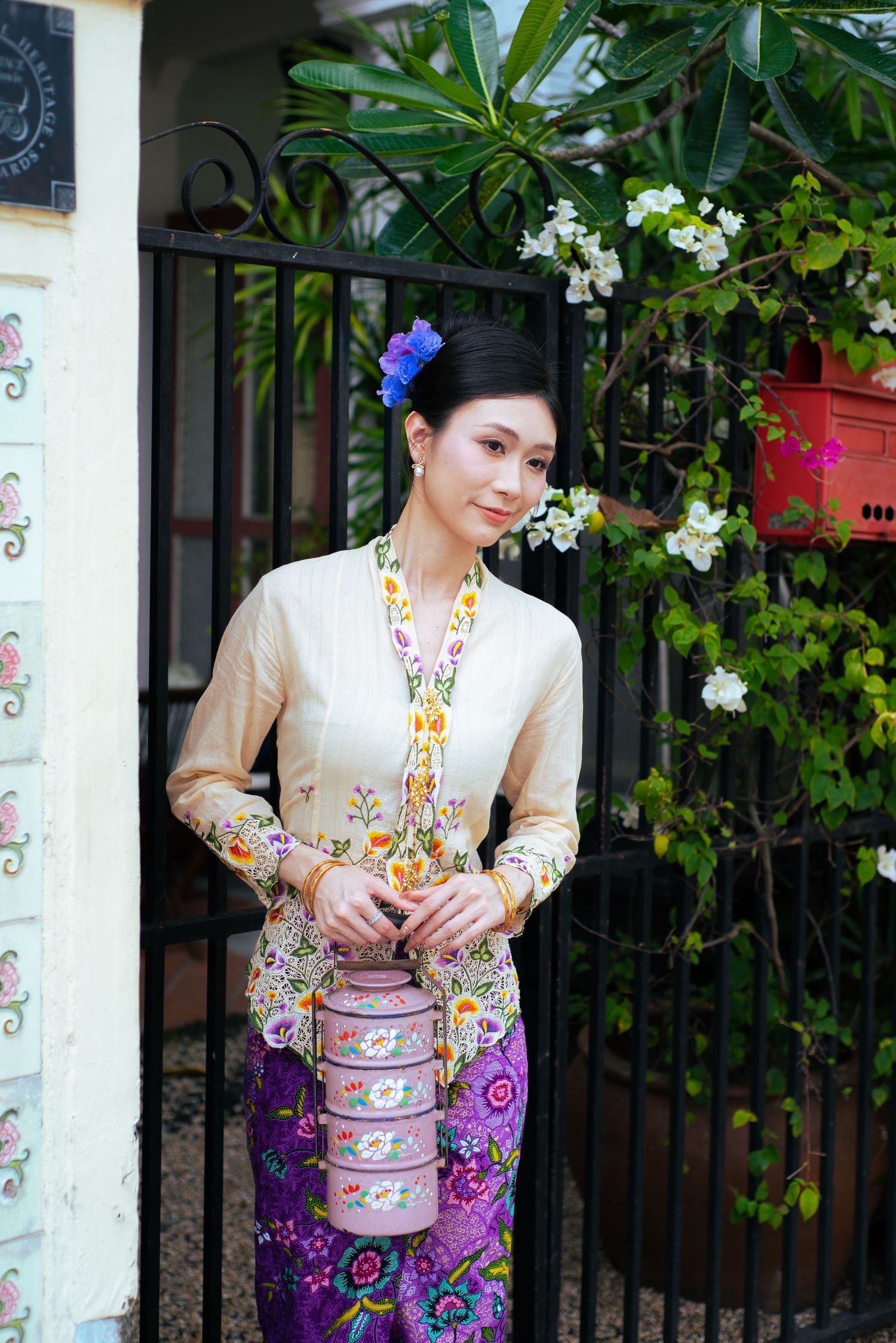 Woman wearing traditional Peranakan Nyonya kebaya dress with beaded necklace and floral motifs stands by iron gate and flowers.
