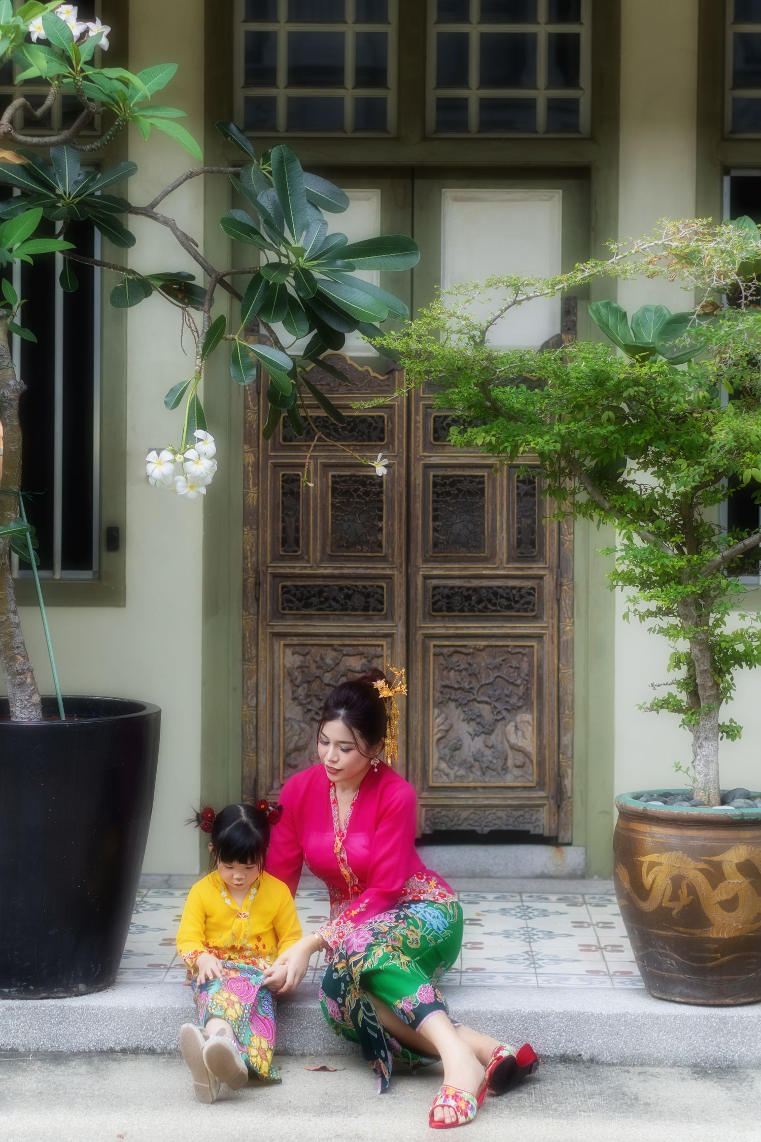 Photo of a Nyonya mother and child in pink and yellow kebaya, in front of a wooden door Pintu pagar, at a Peranakan house in Joo Chiat