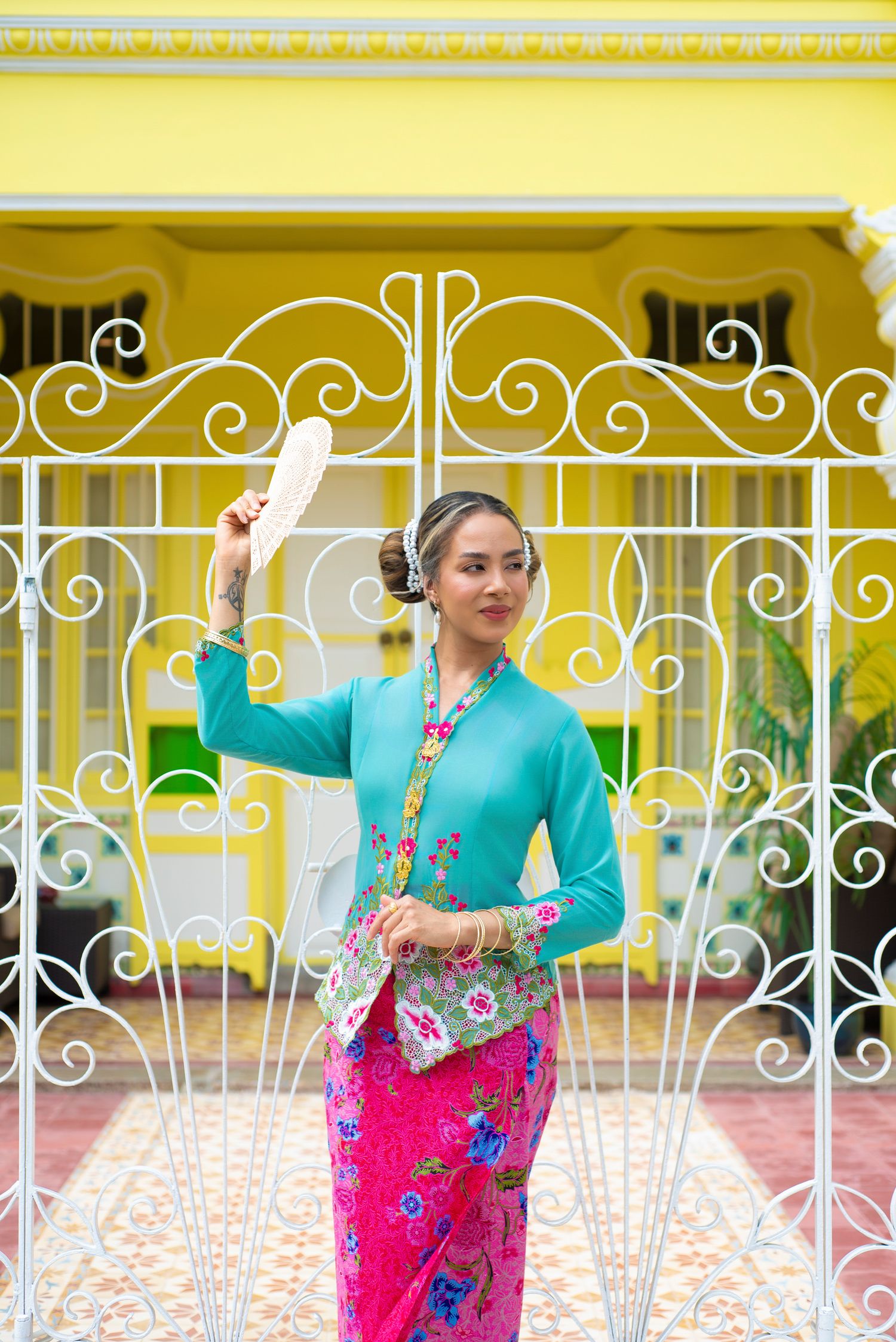 Traditional dancer in turquoise kebaya and pink batik sarong performs graceful movements against ornate white gate.