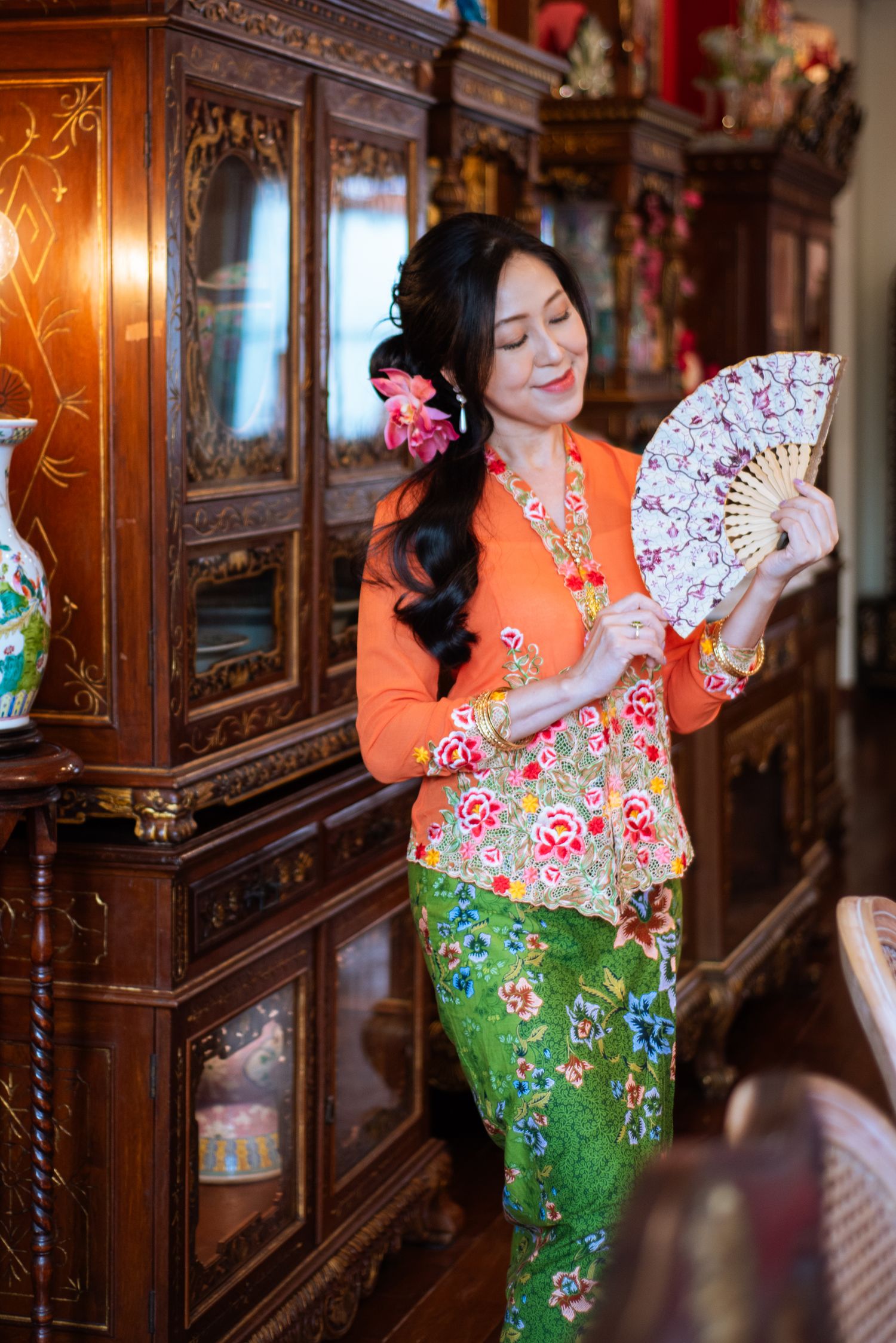 Woman in traditional peranakan kebaya outfit with orange top and green skirt holding decorative fan in antique setting.