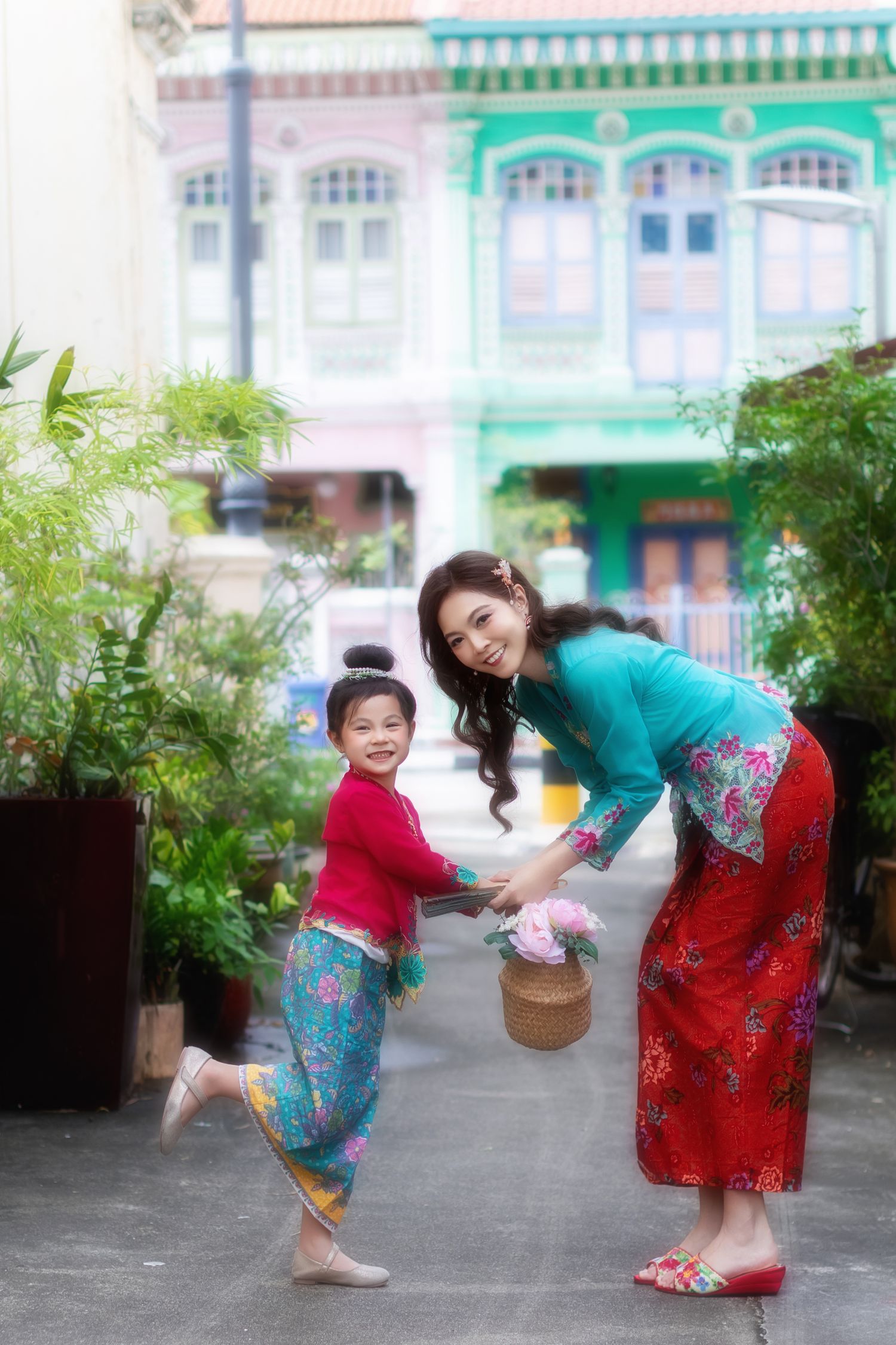 Photo of a mother and daughter in traditional Nyonya Kebaya walking together carrying a basket of flowers, in front of Peranakan houses.