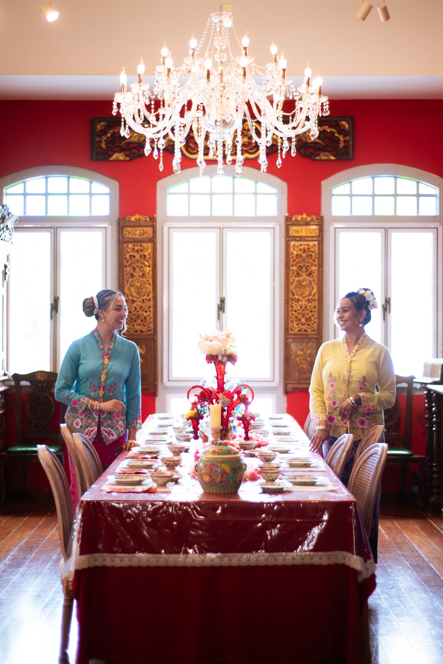 Elegant dining room setup with crystal chandeliers, red walls, and tables dressed in burgundy linens under arched windows.