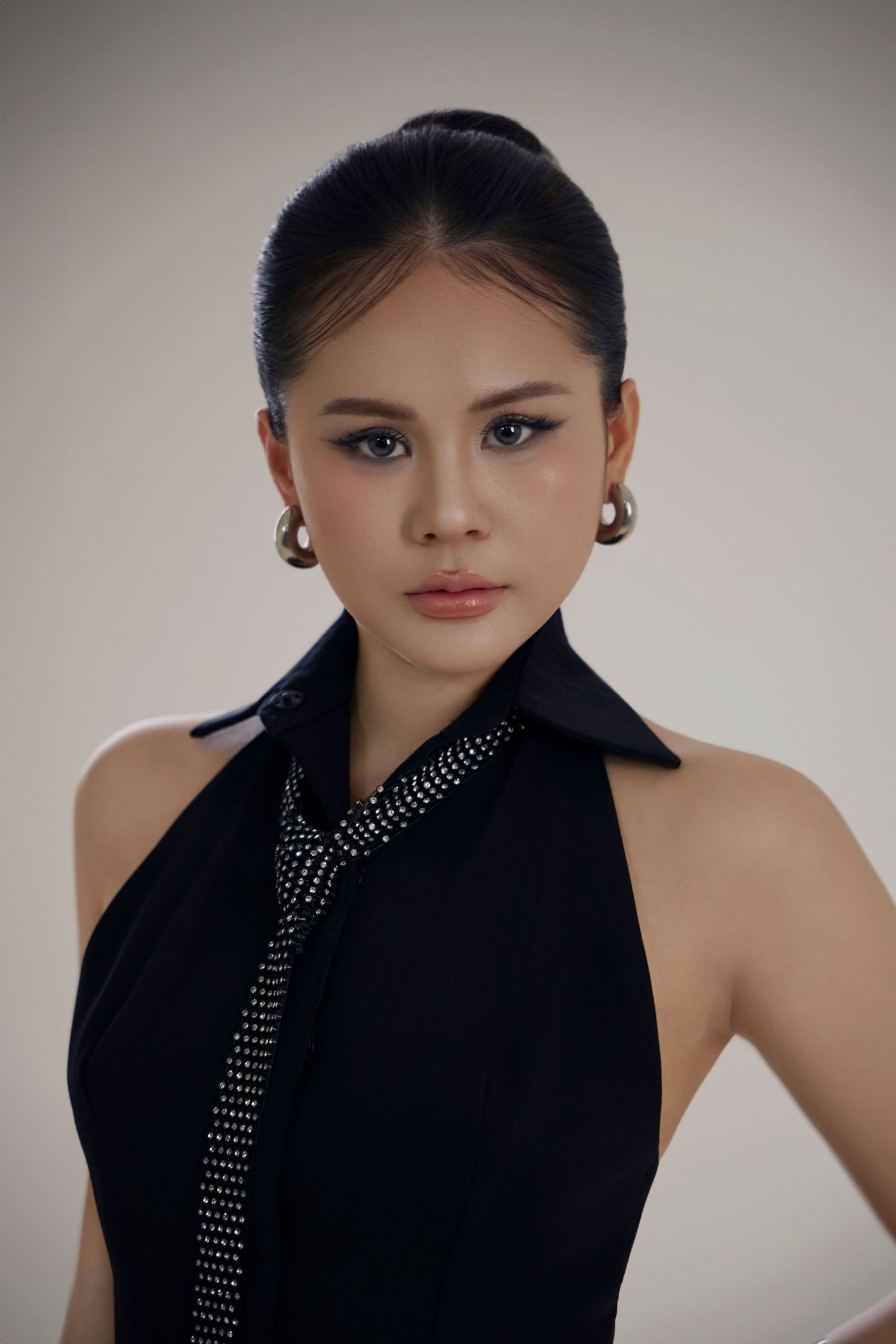 Young Asian woman in black halter top with rhinestone tie, pearl earrings, hair up, posing against neutral background.