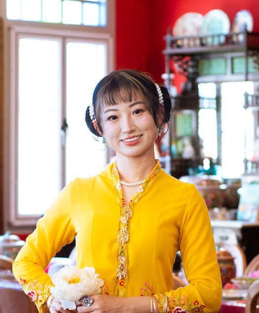 Restaurant staff member wearing a bright yellow sweater and headphones stands in front of a window and display shelves.