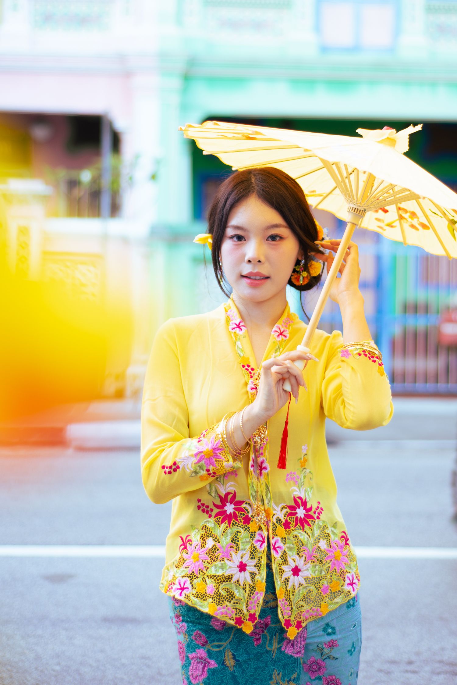 A person in a yellow floral jacket and turquoise dress holds an ornate parasol while walking outdoors.