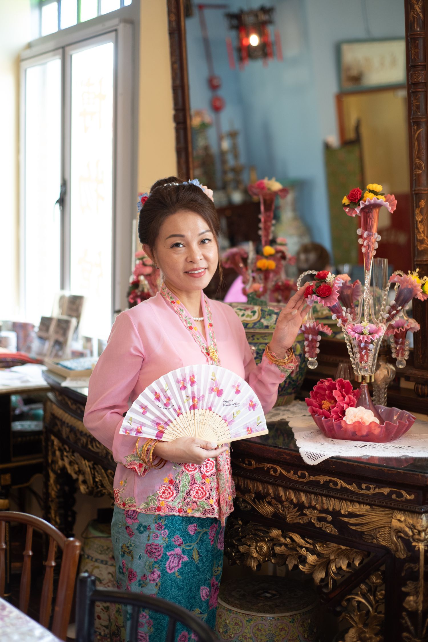 Traditional Peranakan style interior with someone in pink kebaya holding a decorative fan beside ornate table with flowers.