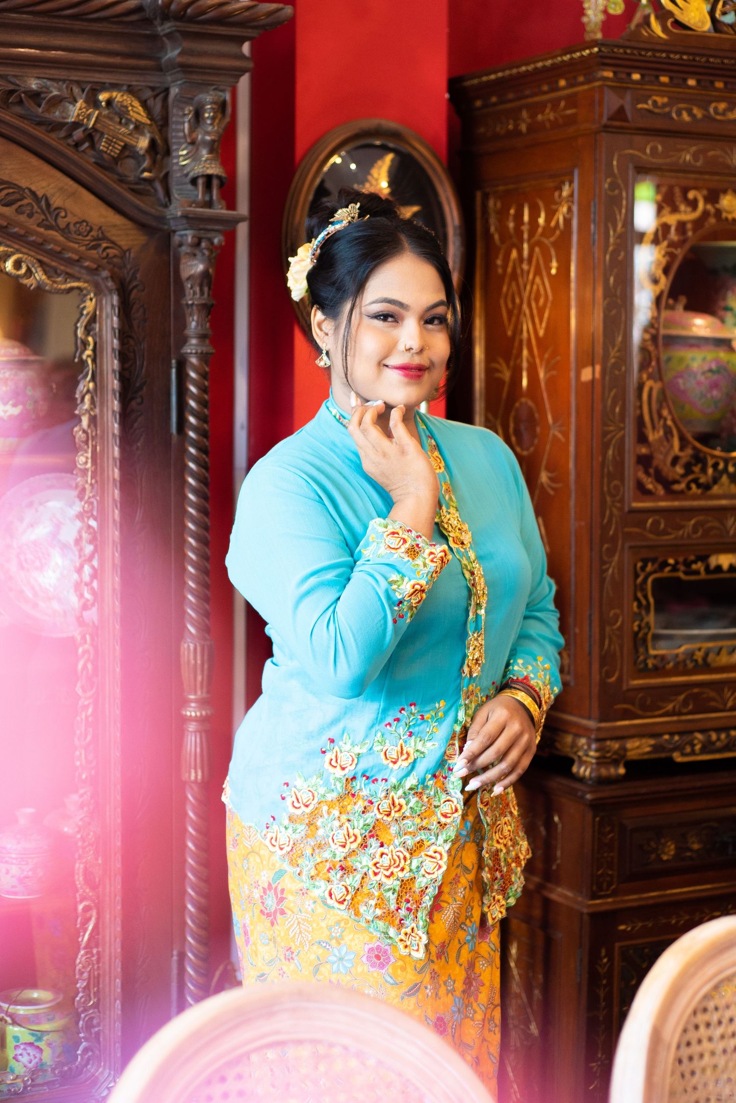 Traditional Southeast Asian dress in turquoise and gold with ornate embellishments, shown against antique carved wooden cabinets.