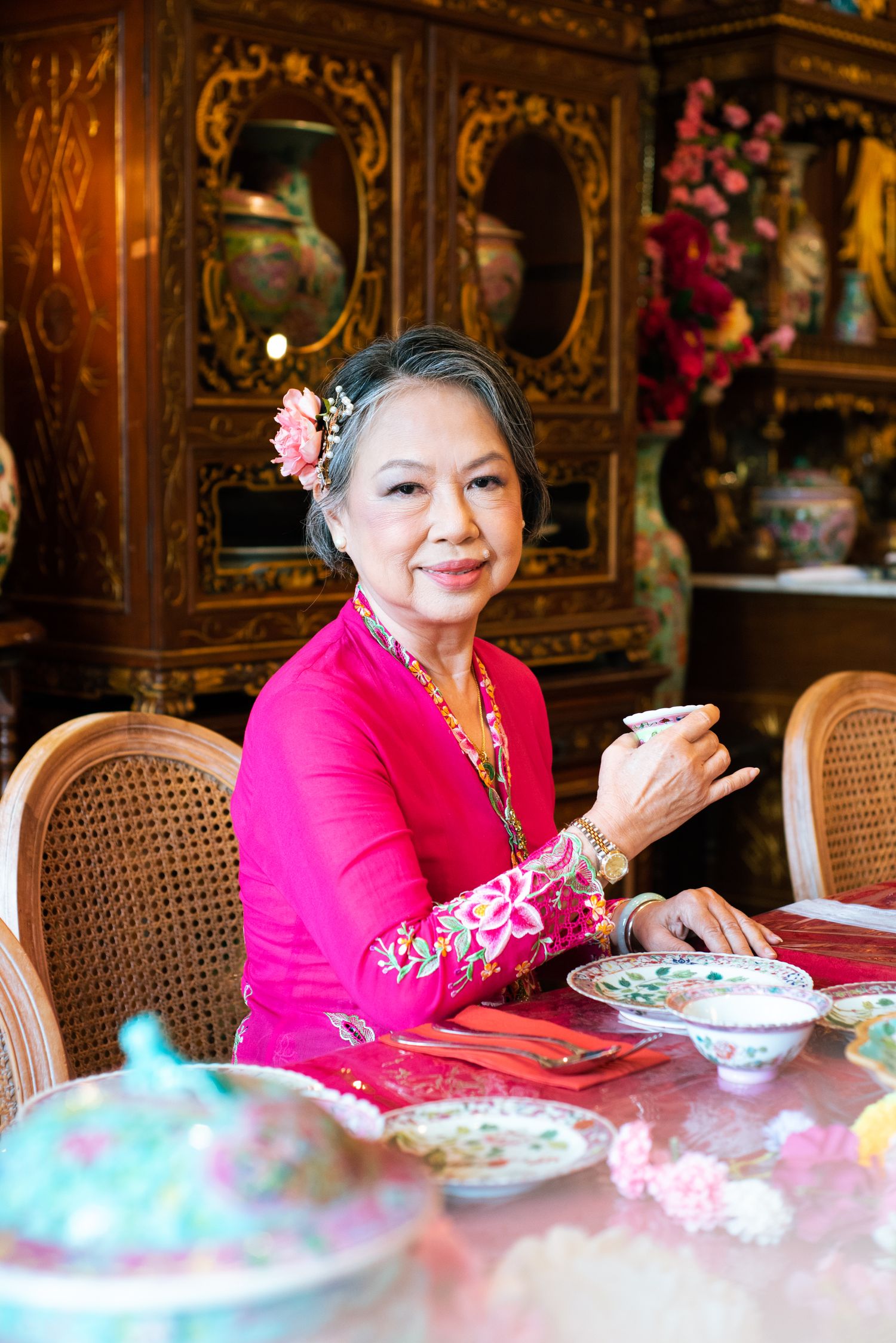 Traditional Asian interior with senior woman in bright pink dress drinking tea at ornate wooden table with tea set.