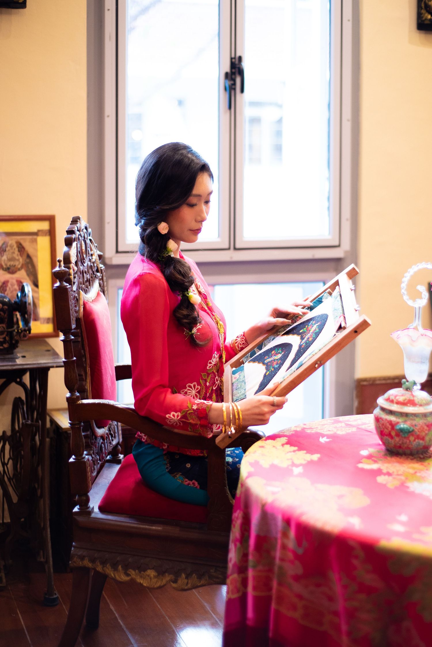 Seated figure in a bright red traditional outfit reads while sitting at a table with a pink tablecloth near a window.