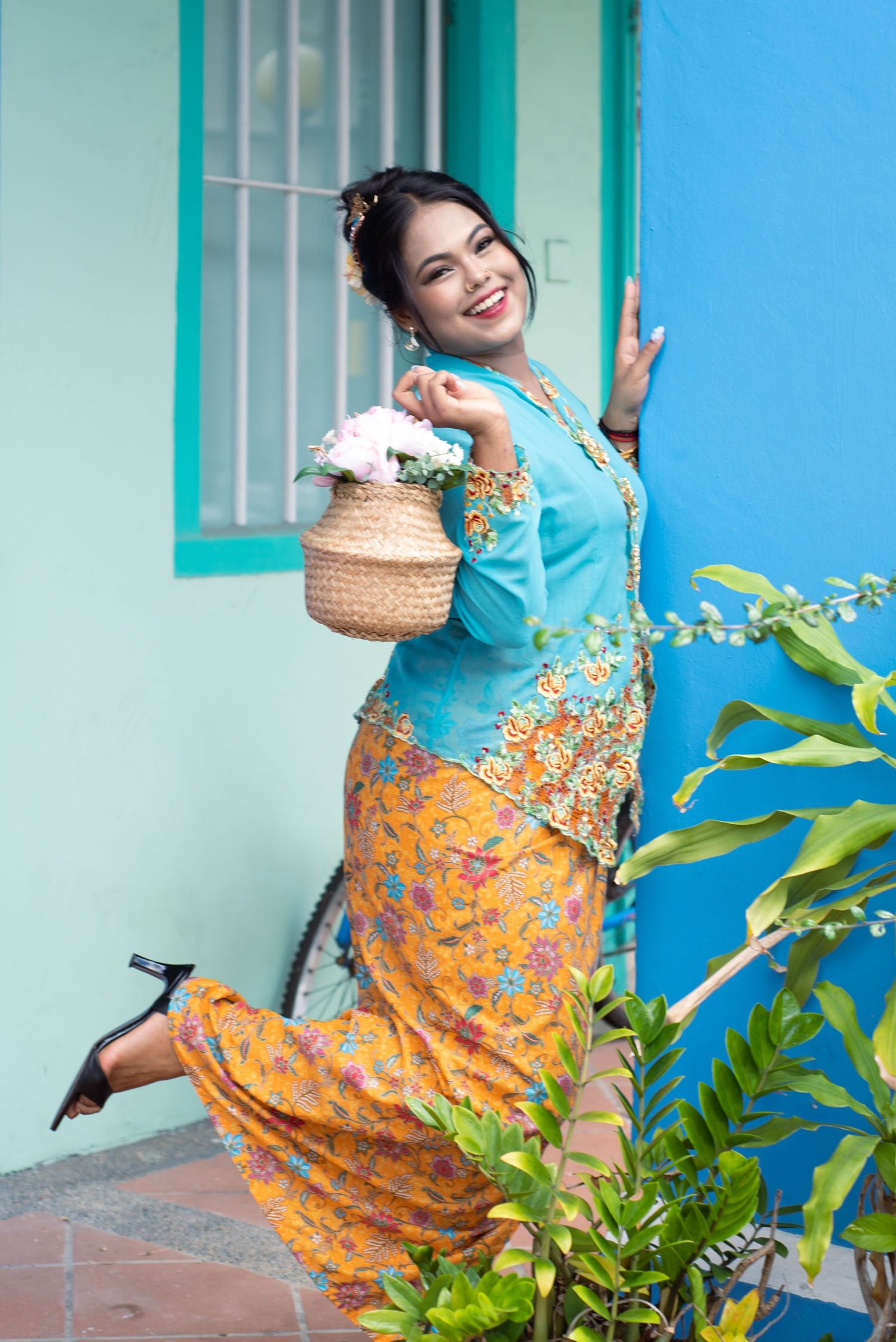 A woman in traditional Peranakan Nyonya kebaya dress poses joyfully with a woven basket against a turquoise wall.