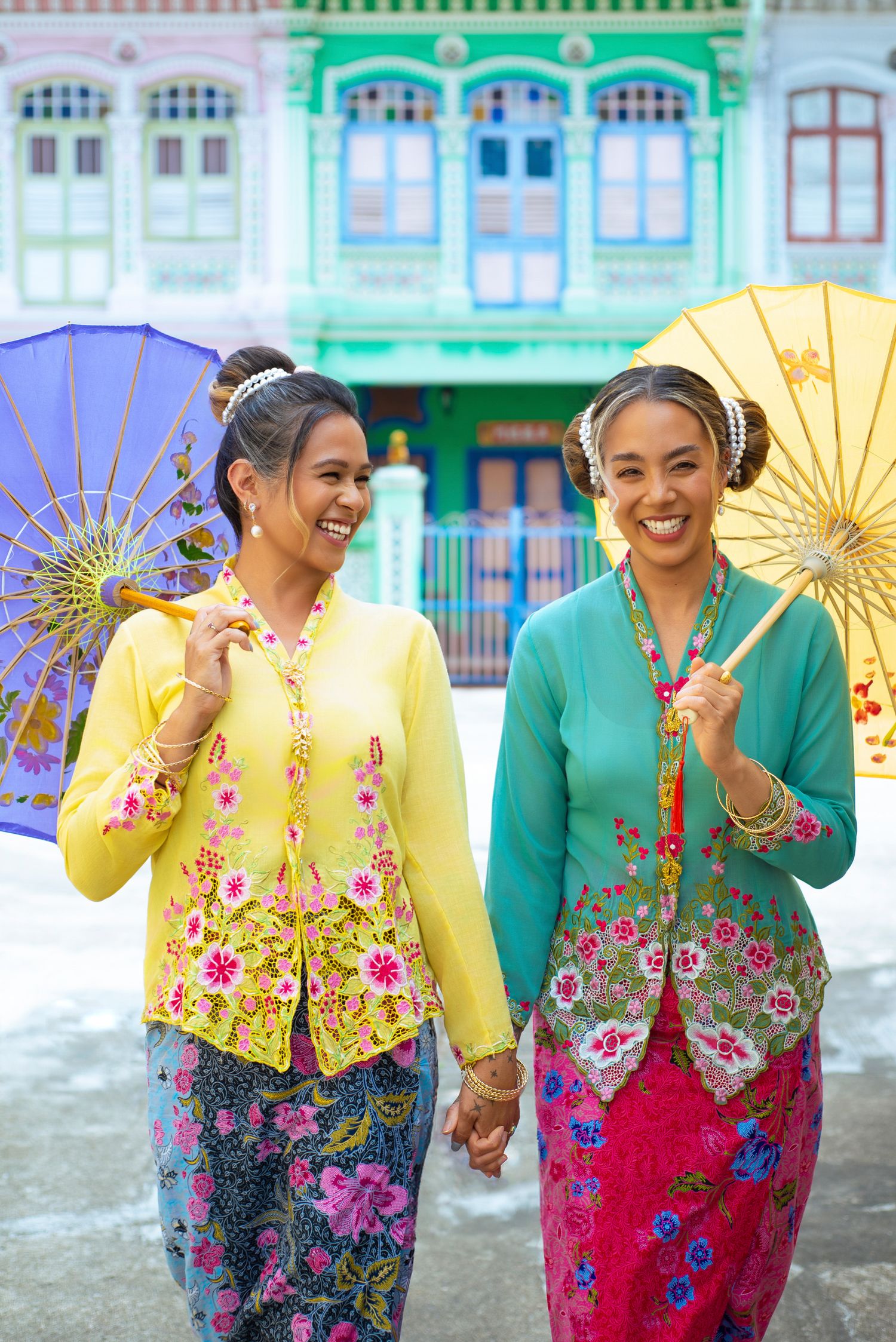 Two people in colorful traditional Peranakan kebaya outfits hold parasols while standing in front of heritage shophouses.