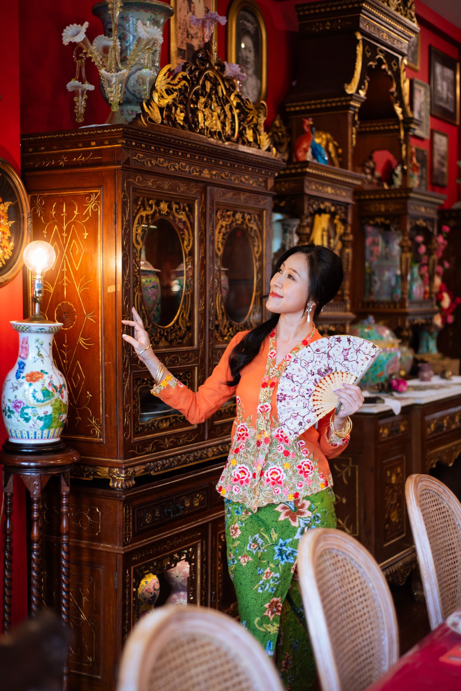 A person in traditional Asian attire poses with ornate porcelain vases in a richly decorated antique room.