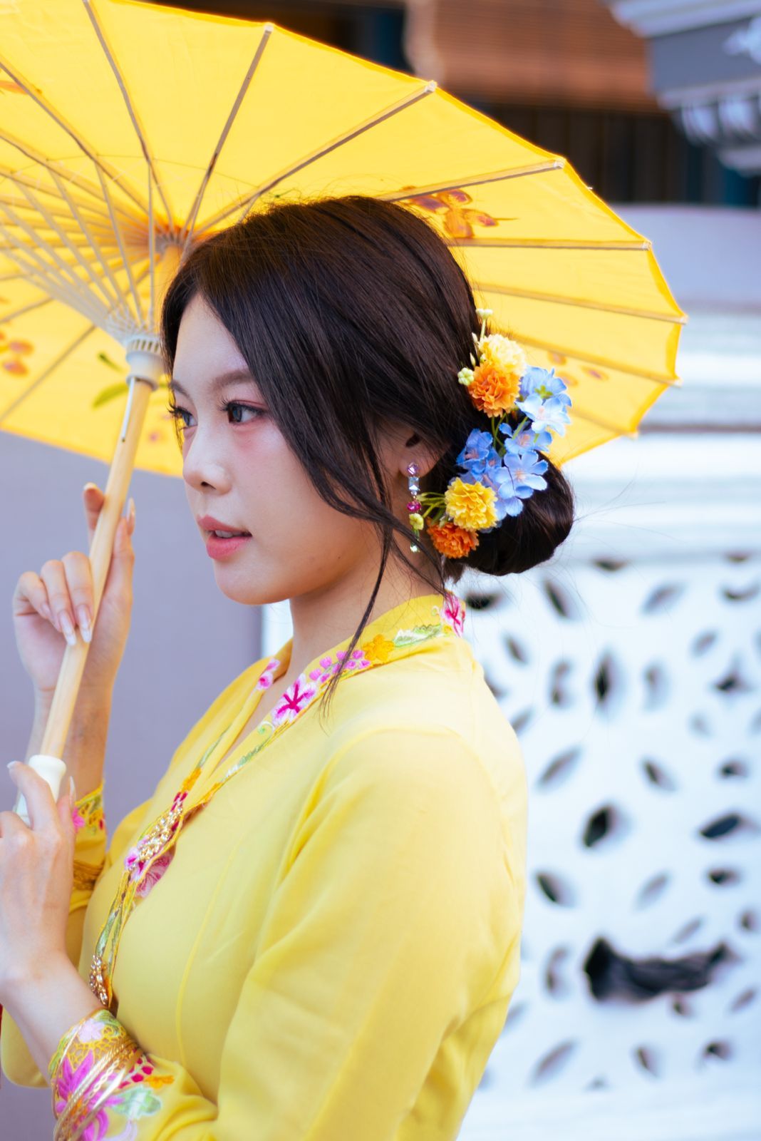 A person in a yellow outfit holds a traditional yellow paper parasol while wearing blue and orange flowers in their hair.