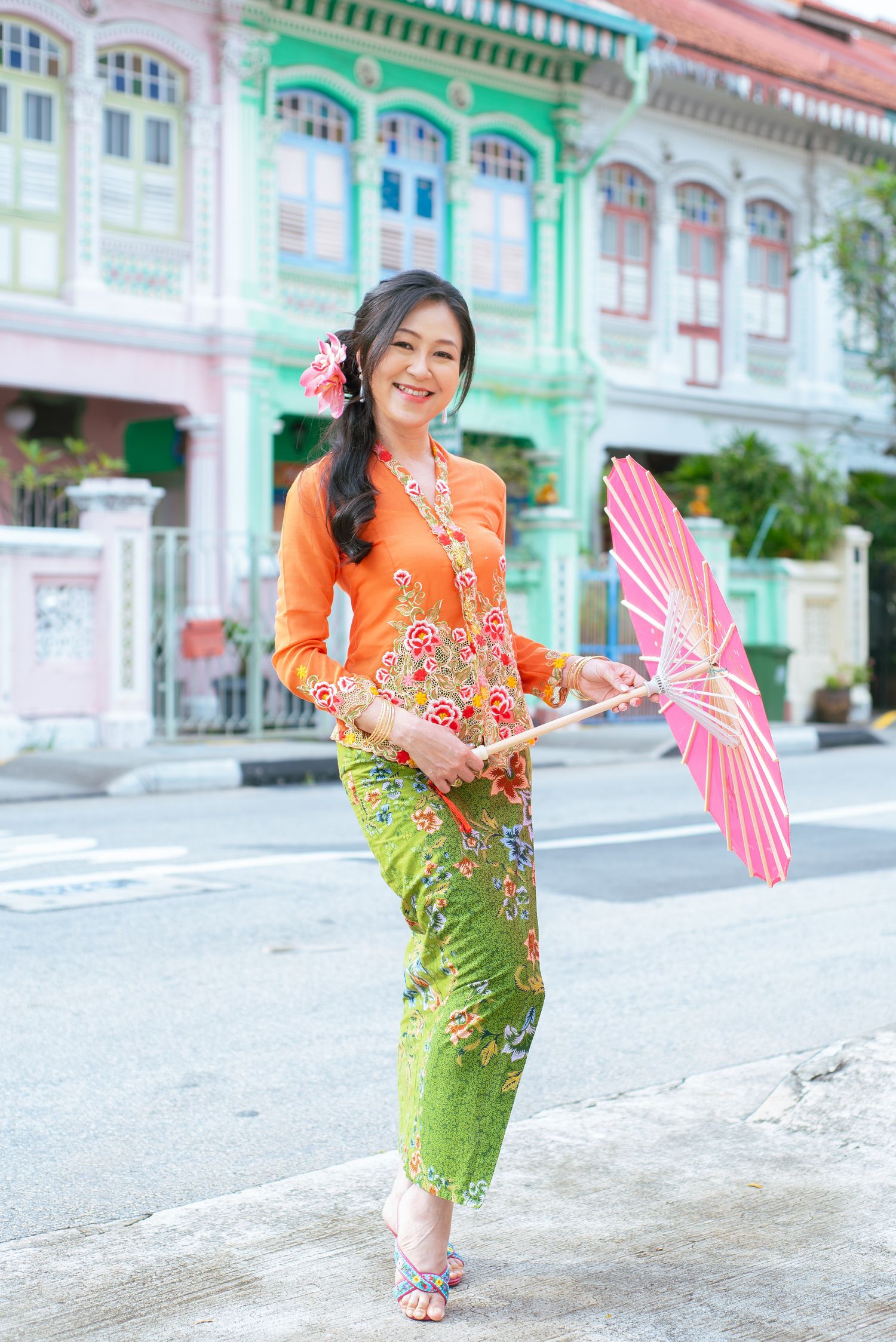 Traditional Peranakan outfit with orange kebaya top and green sarong skirt holding pink parasol in front of heritage shophouses.