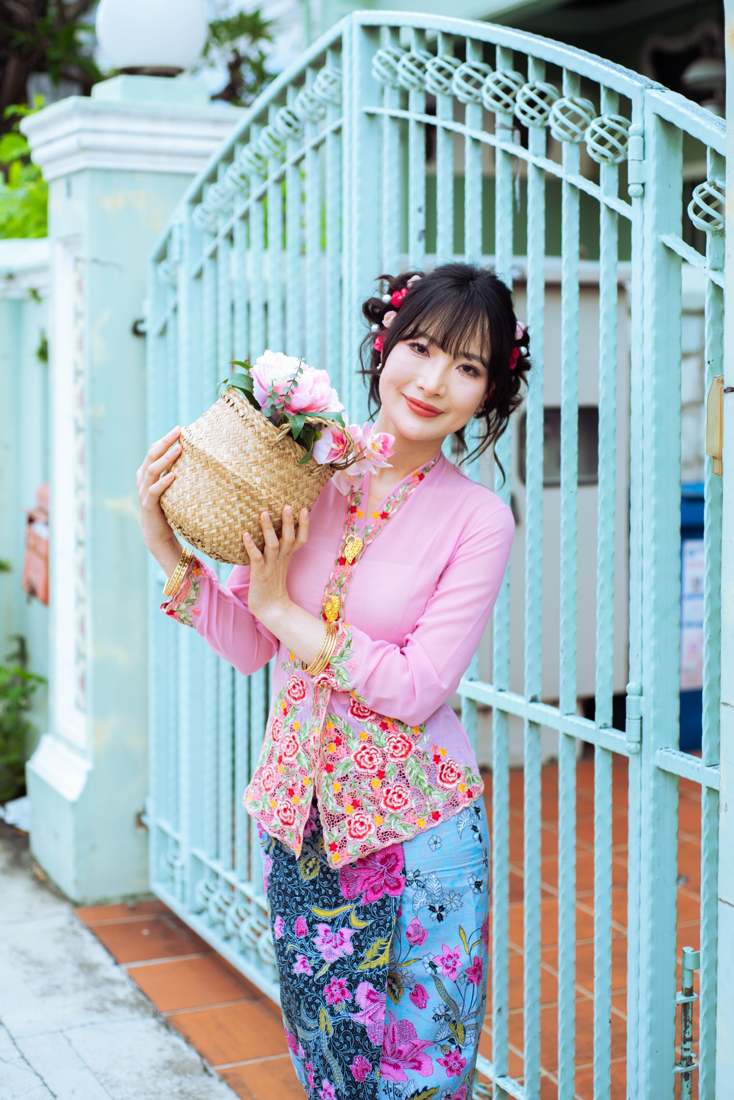Someone in pink floral traditional clothing holds a wicker basket with flowers by an ornate blue gate.