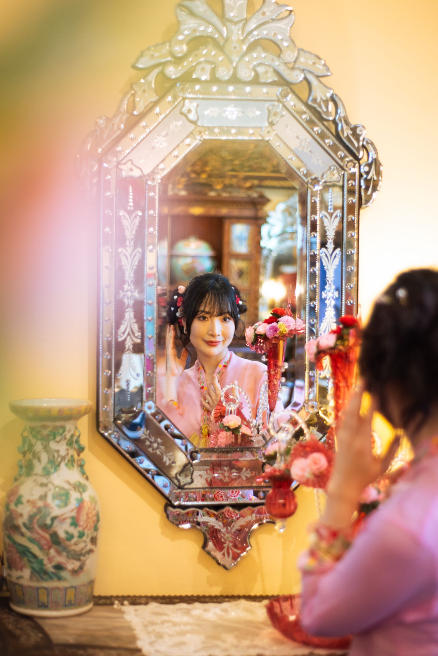 A decorative Venetian-style mirror with etched glass details hangs on a yellow wall beside an ornate vase.