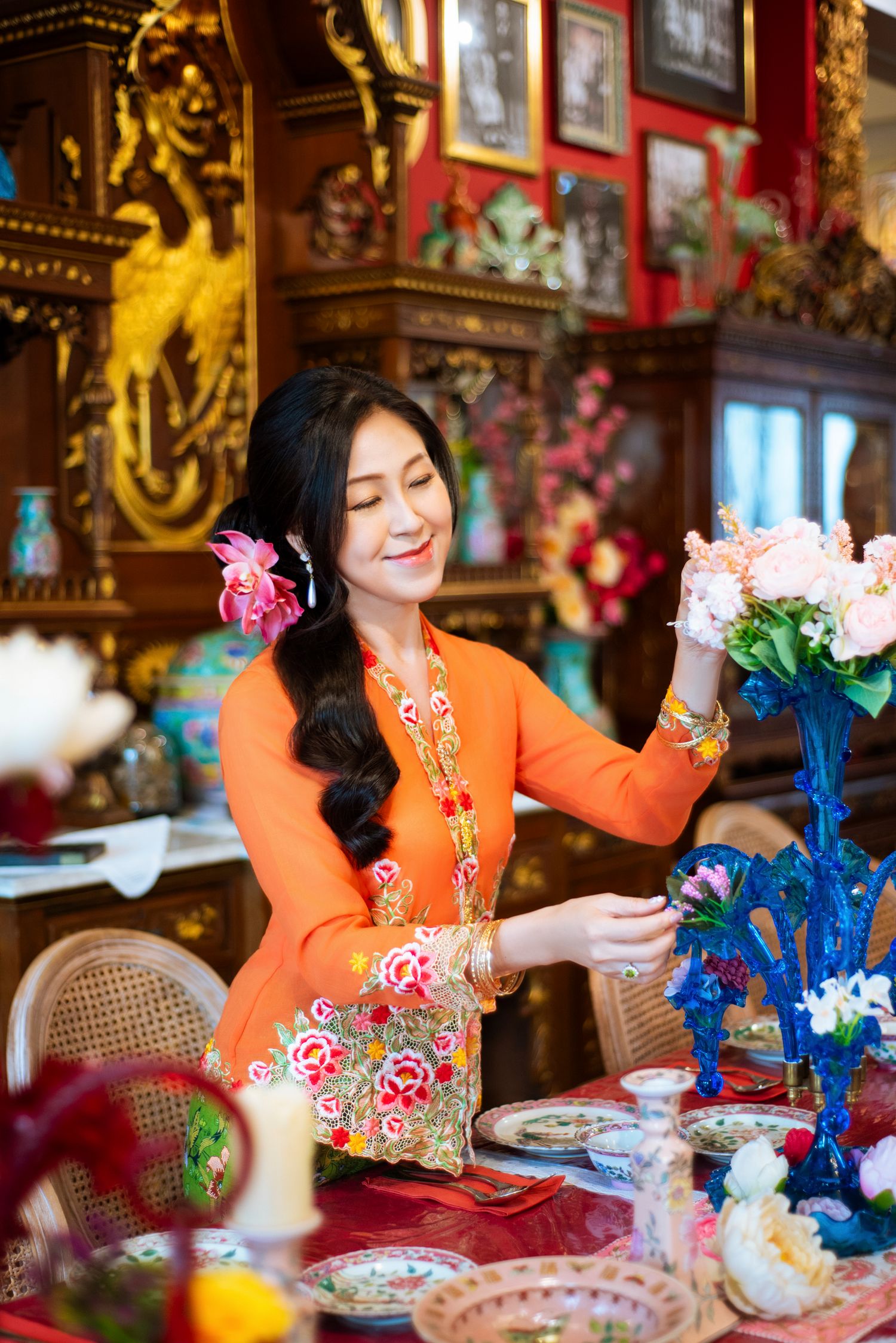 Person in bright orange dress dines at ornate table decorated with pink and blue flowers in elegant room setting.
