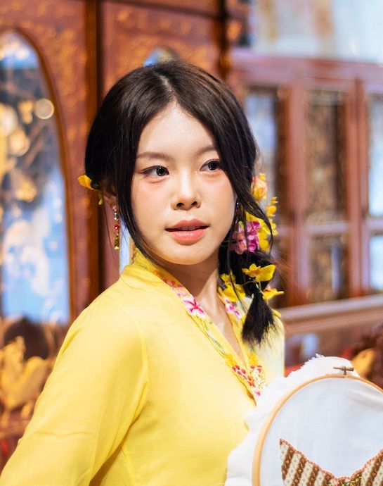 Woman in bright yellow sweater sitting with colorful decorative elements in warm indoor setting with bokeh lights.