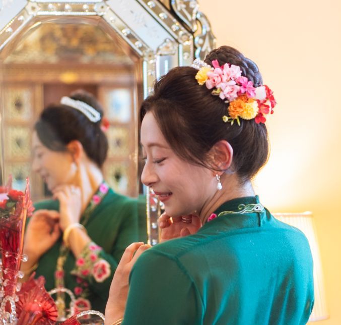 Person wearing a green traditional dress with colorful floral hair accessories smiles during a celebration.