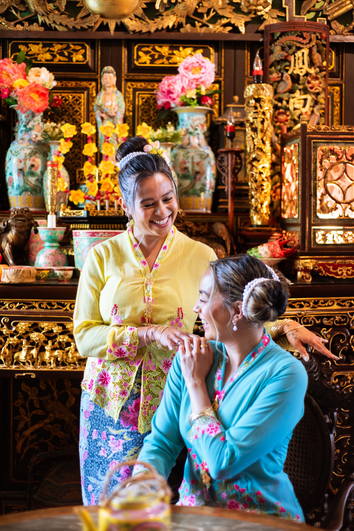 Two people wearing colorful traditional Peranakan outfits interact in front of an ornate altar with flowers and decorations.