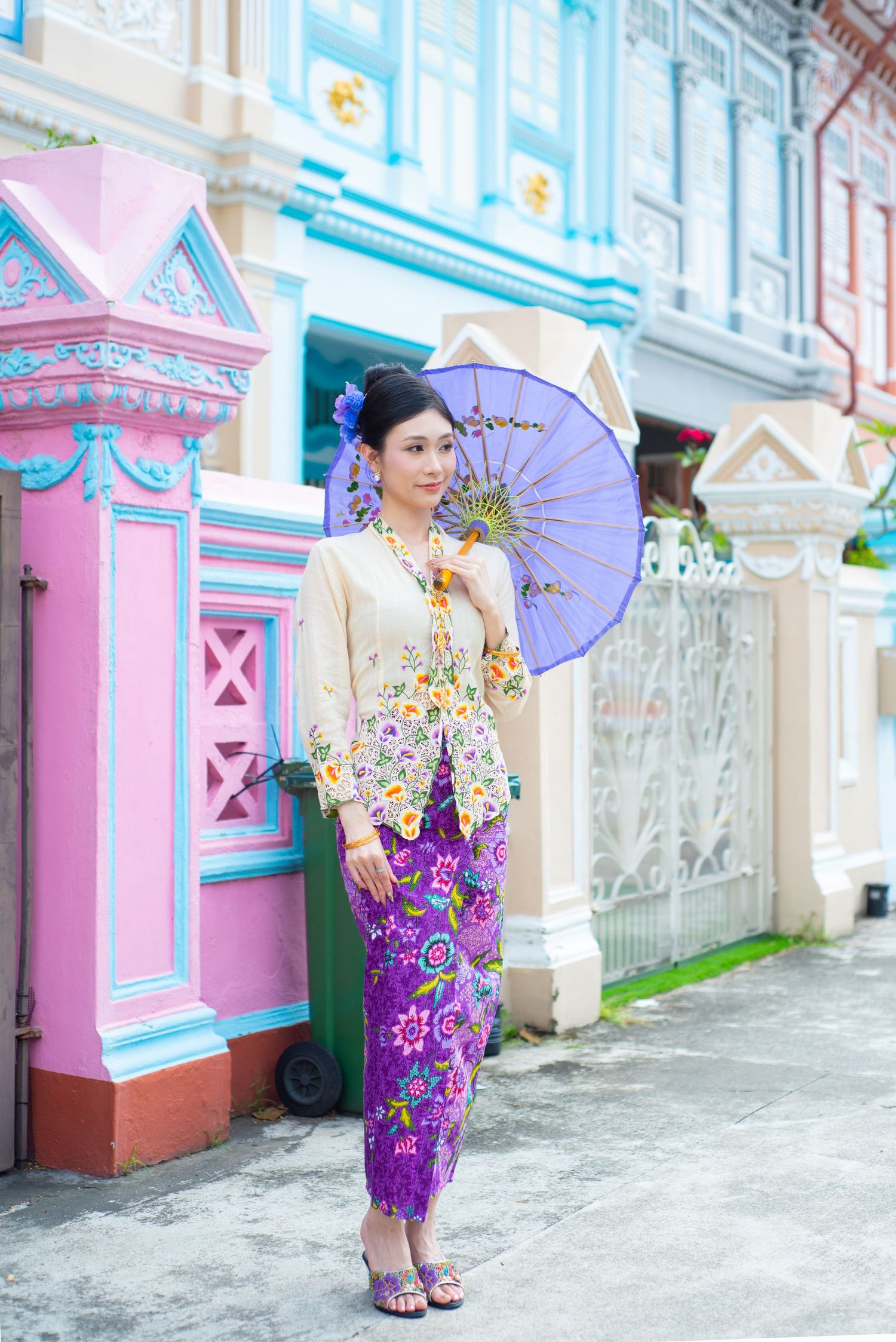 Traditional Thai formal attire worn with a purple parasol against a colorful pastel building with ornate architectural details.